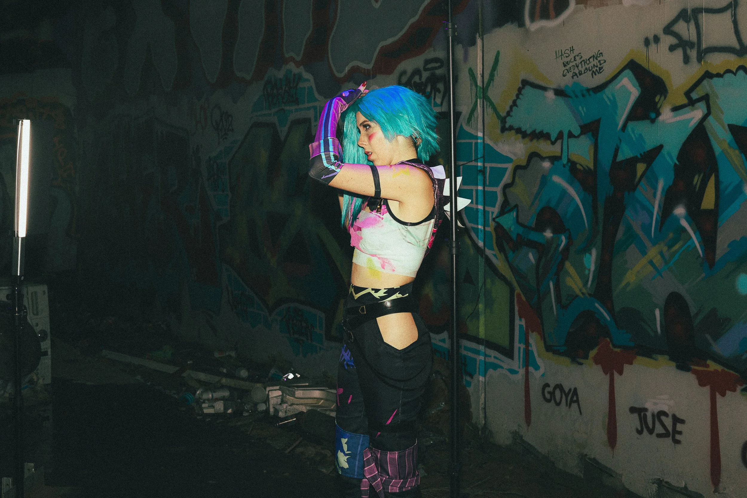 A woman with blue hair and colorful clothing in an urban graffiti-covered room, adjusting her hair.