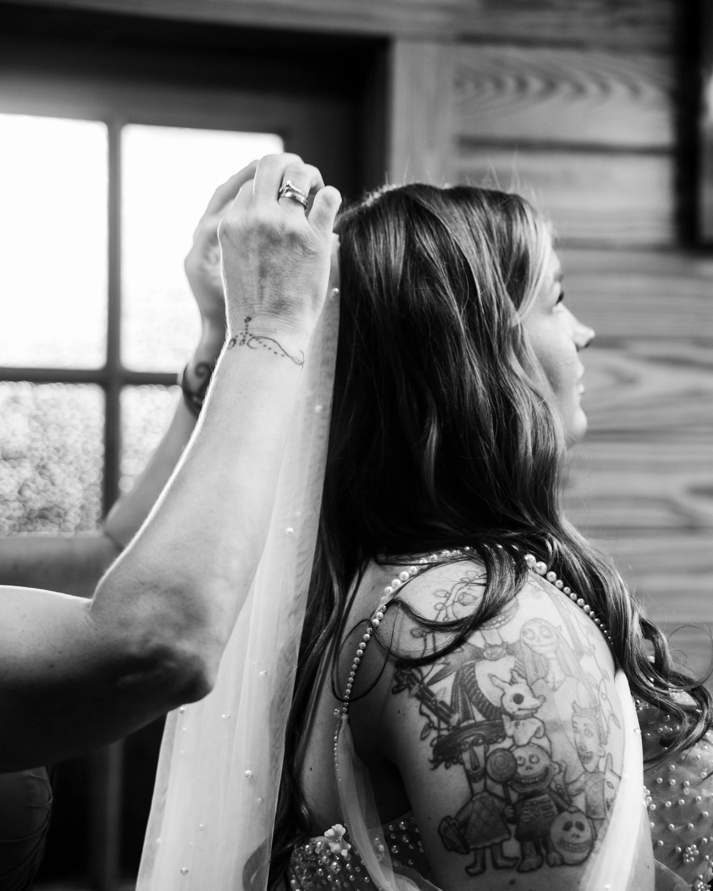 Woman with tattooed arm and back, getting her veil arranged, near a window with natural light.