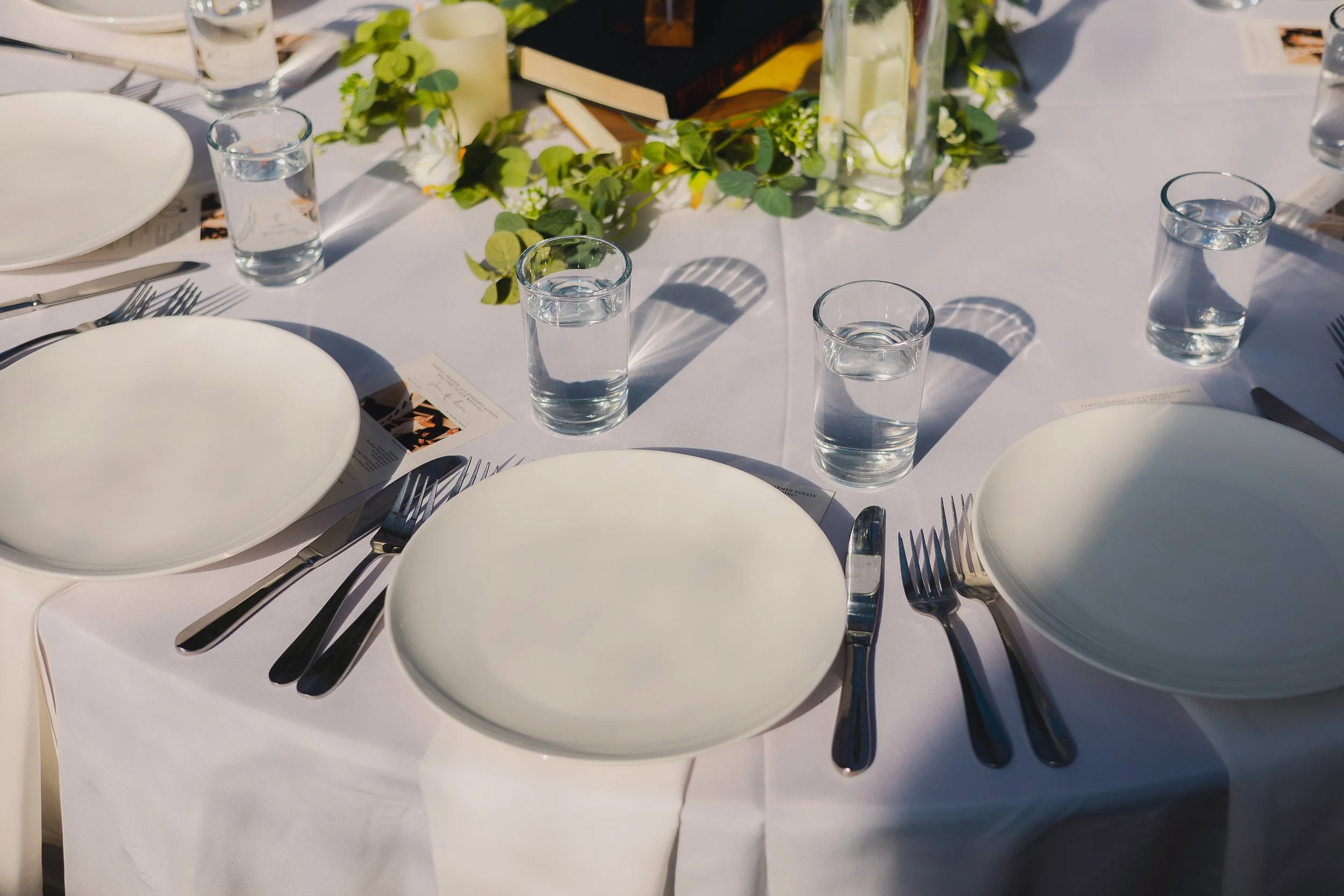 A formal dining table set with three white plates, silver cutlery, and four glasses of water. The table has a white tablecloth and a decorative centerpiece with greenery and glass vases with lemon slices.