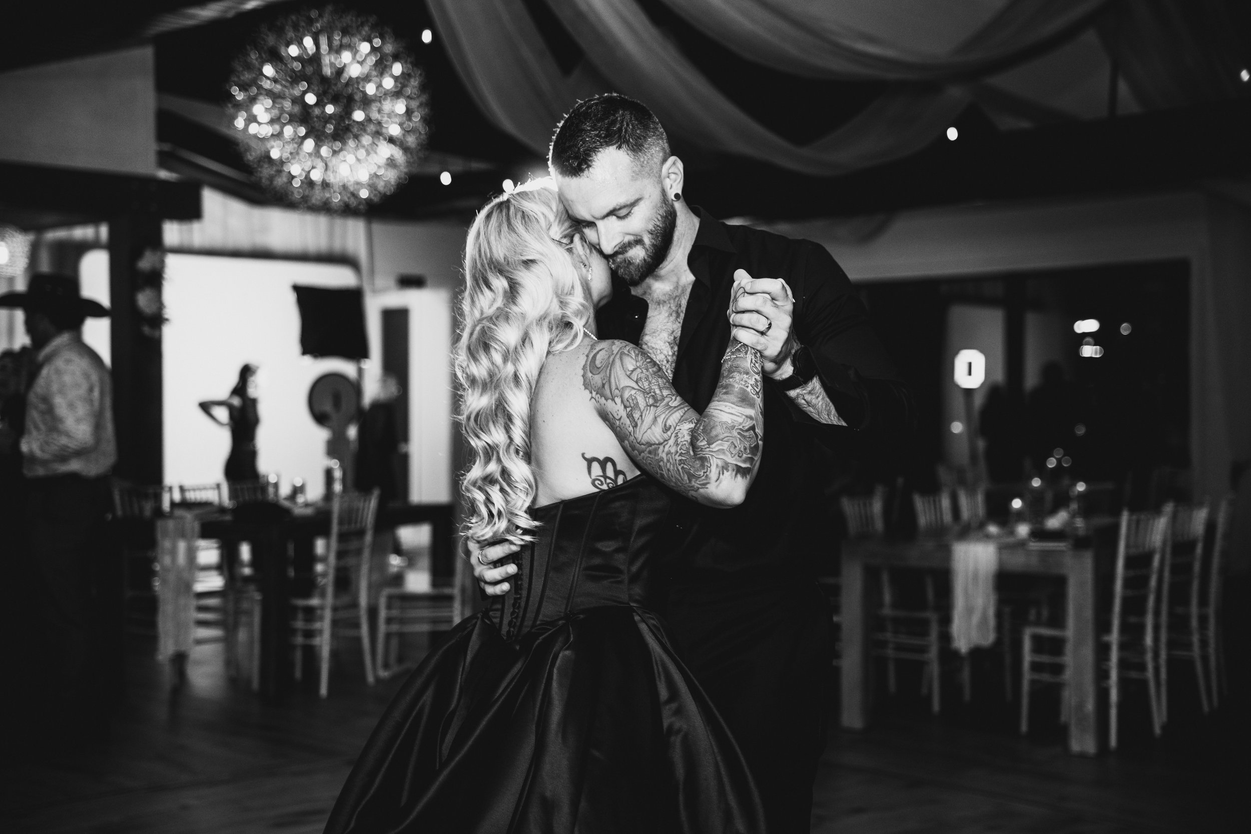 A man and a woman dancing closely at a dimly lit event venue. The woman has long wavy hair and visible tattoos, and the man has short hair and a beard. They are holding hands and appear to be sharing an intimate moment.