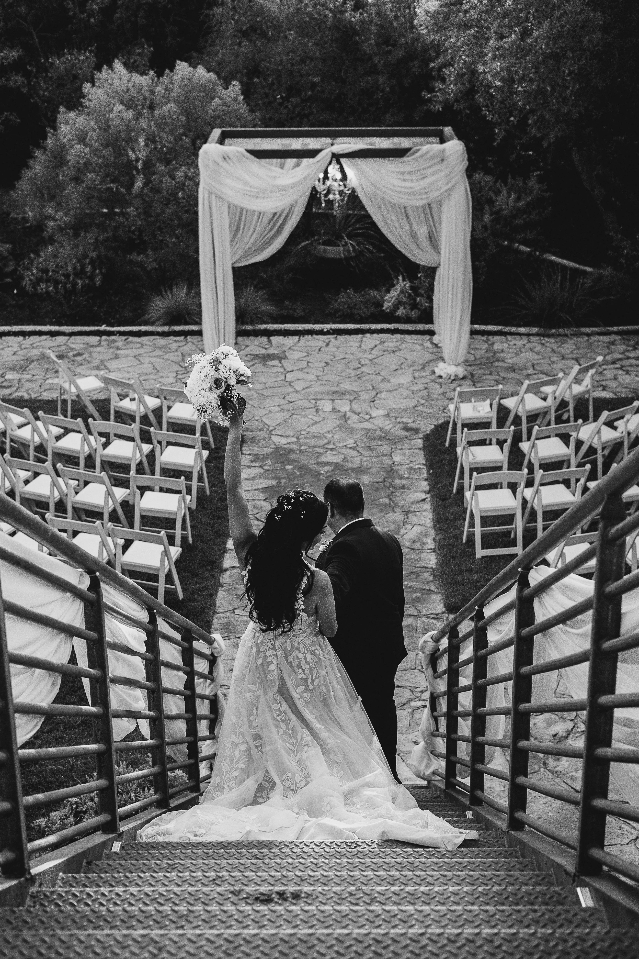 Black and white photo of a bride and groom standing on a staircase, the bride is holding a bouquet with one arm raised, and the wedding ceremony setup includes chairs and an altar with drapery and a chandelier in the background.