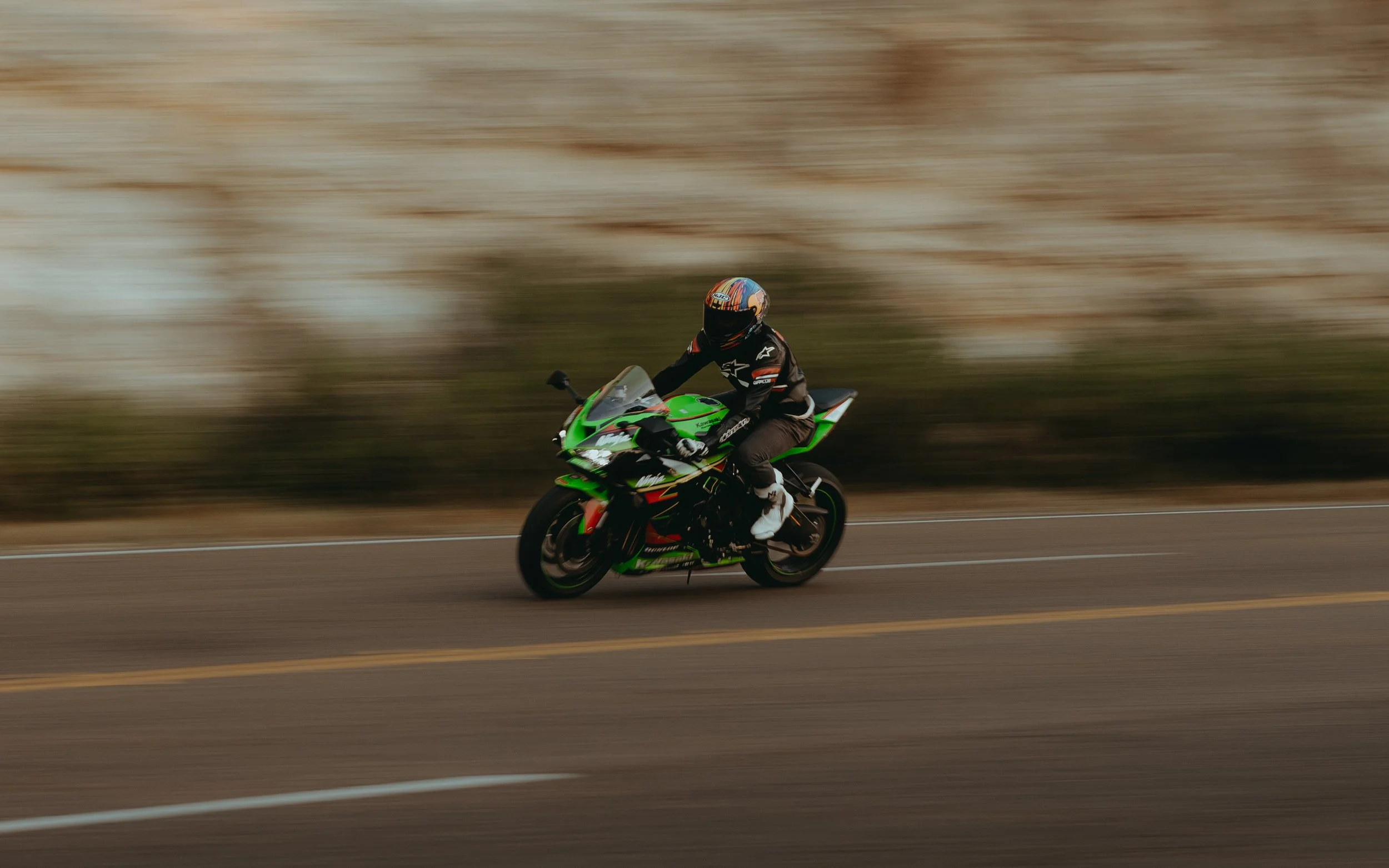 A motorcyclist wearing a black and white racing suit and a colorful helmet riding a green Kawasaki sportbike on a paved road with a blurred desert landscape background.