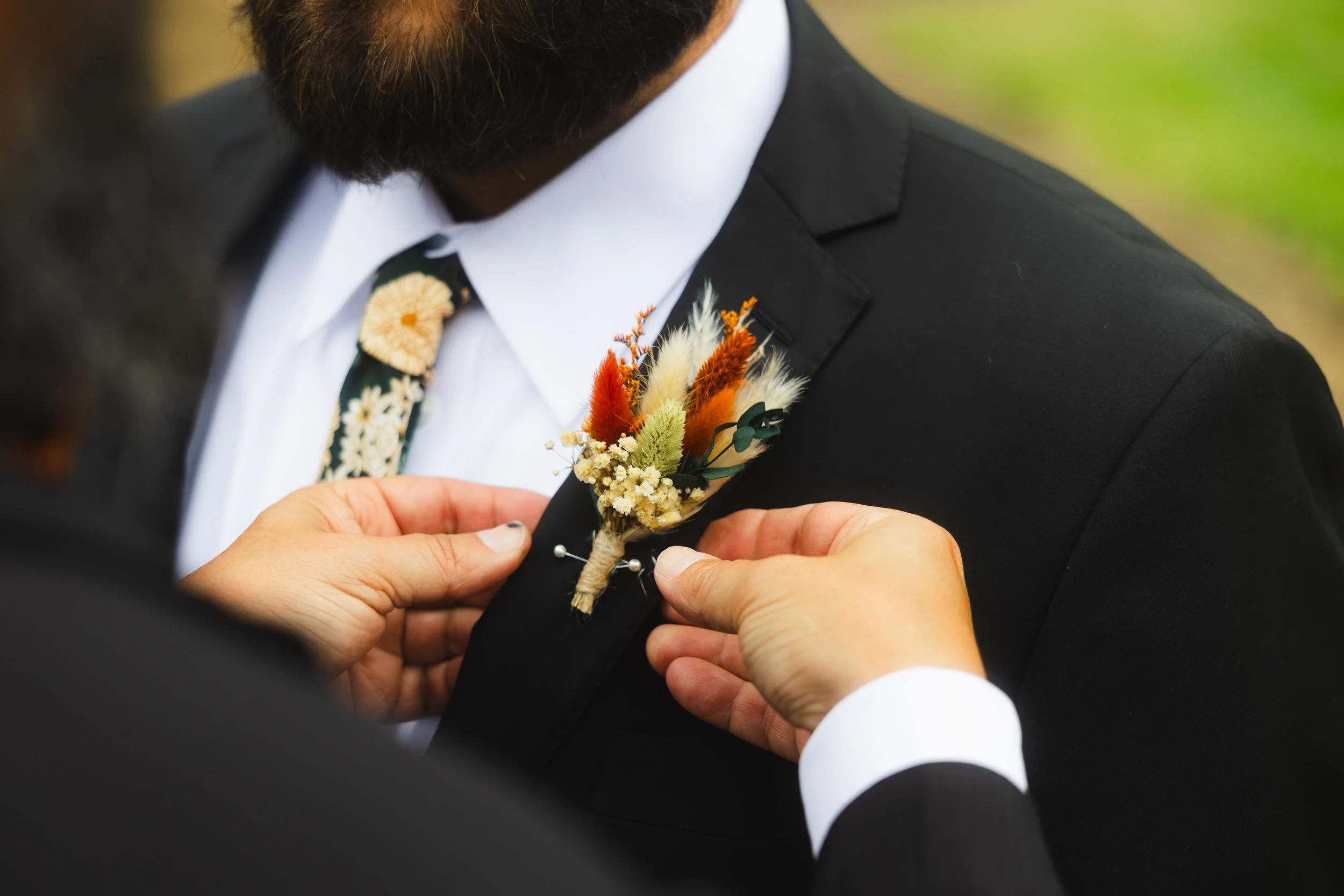 Person pinning a boutonniere with dried flowers to a man's suit jacket during a formal event.