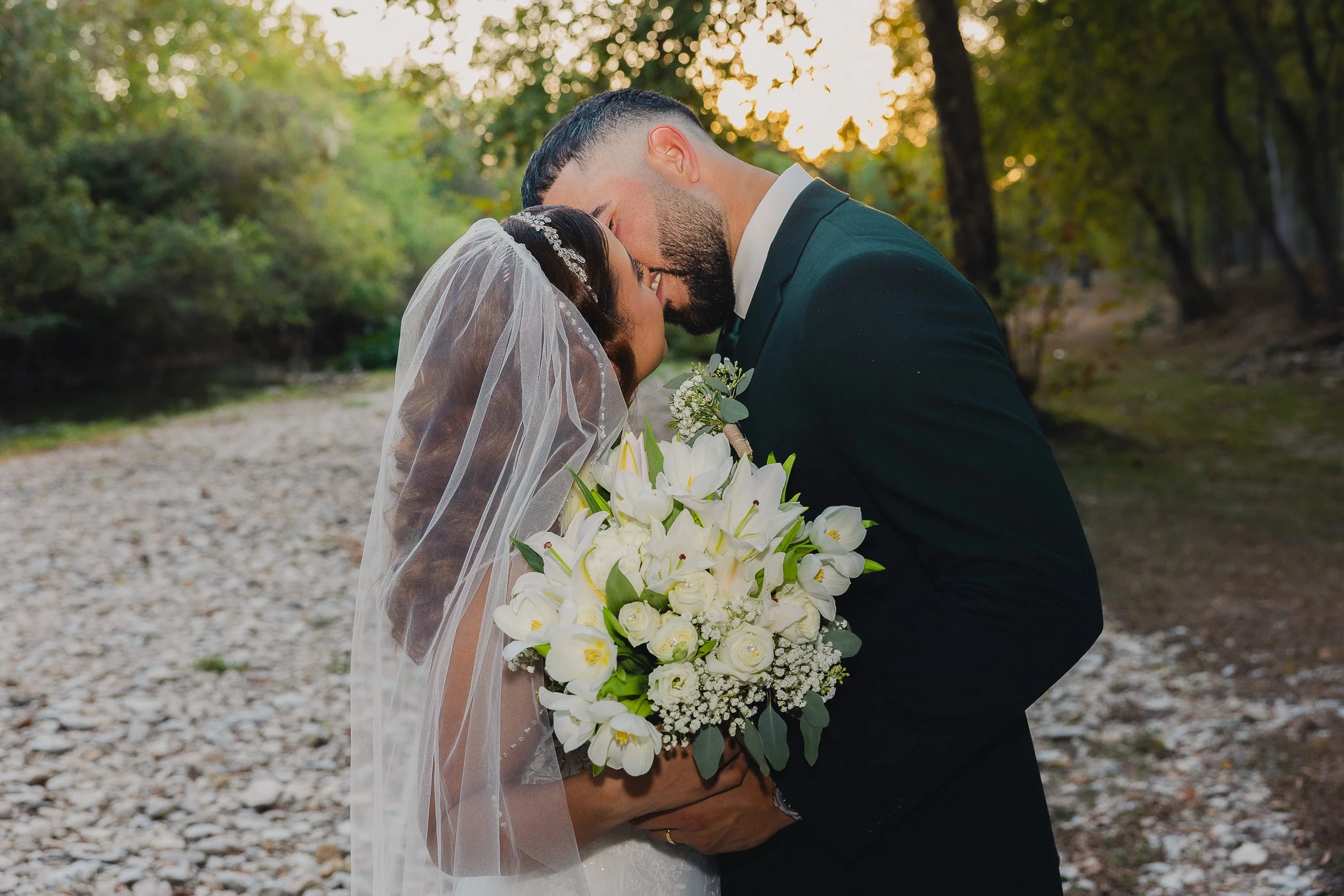 A bride and groom sharing a kiss outdoors during sunset, with the bride holding a large bouquet of white flowers, surrounded by trees and a rocky path.