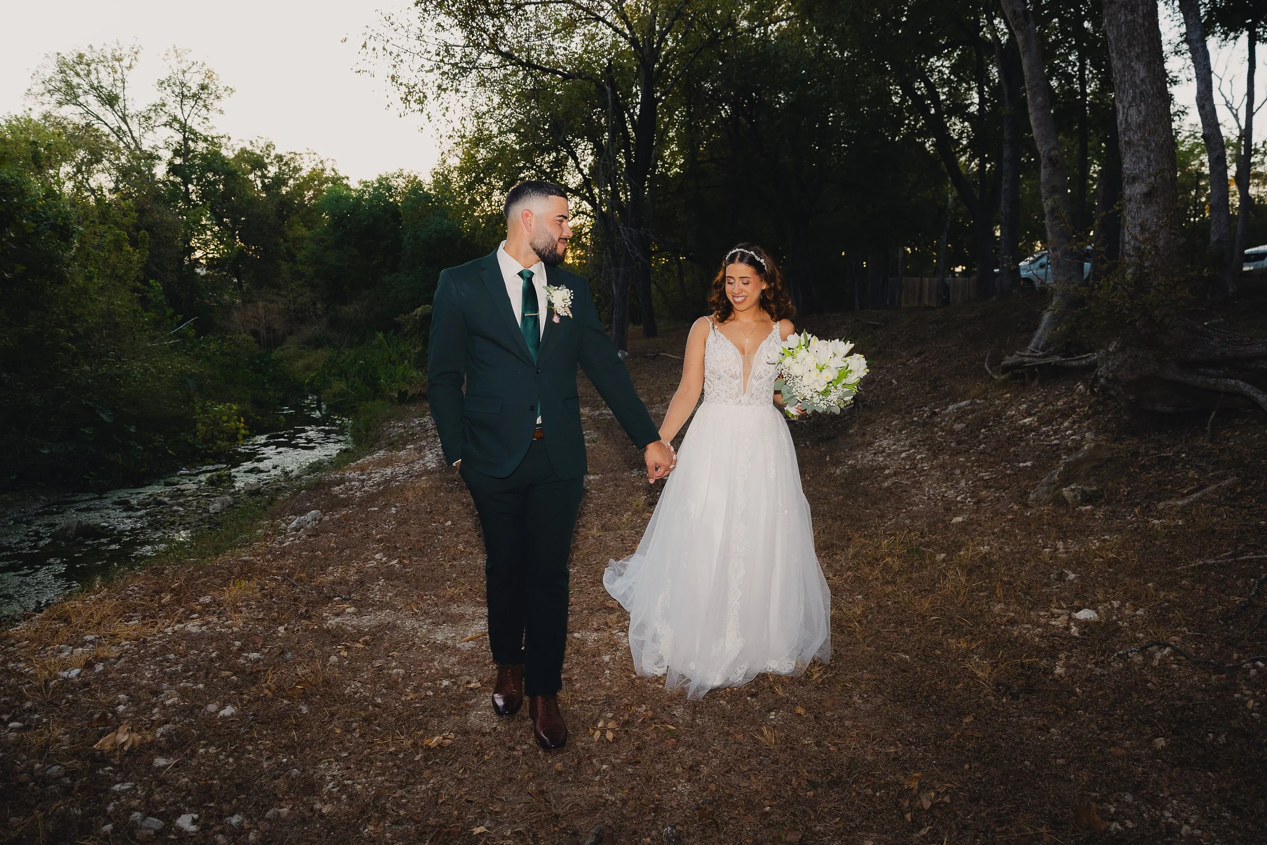 A bride and groom walking hand-in-hand along a wooded nature trail during their wedding, with the bride holding a bouquet of white flowers and wearing a white lace wedding dress, while the groom is in a dark suit with a green tie.