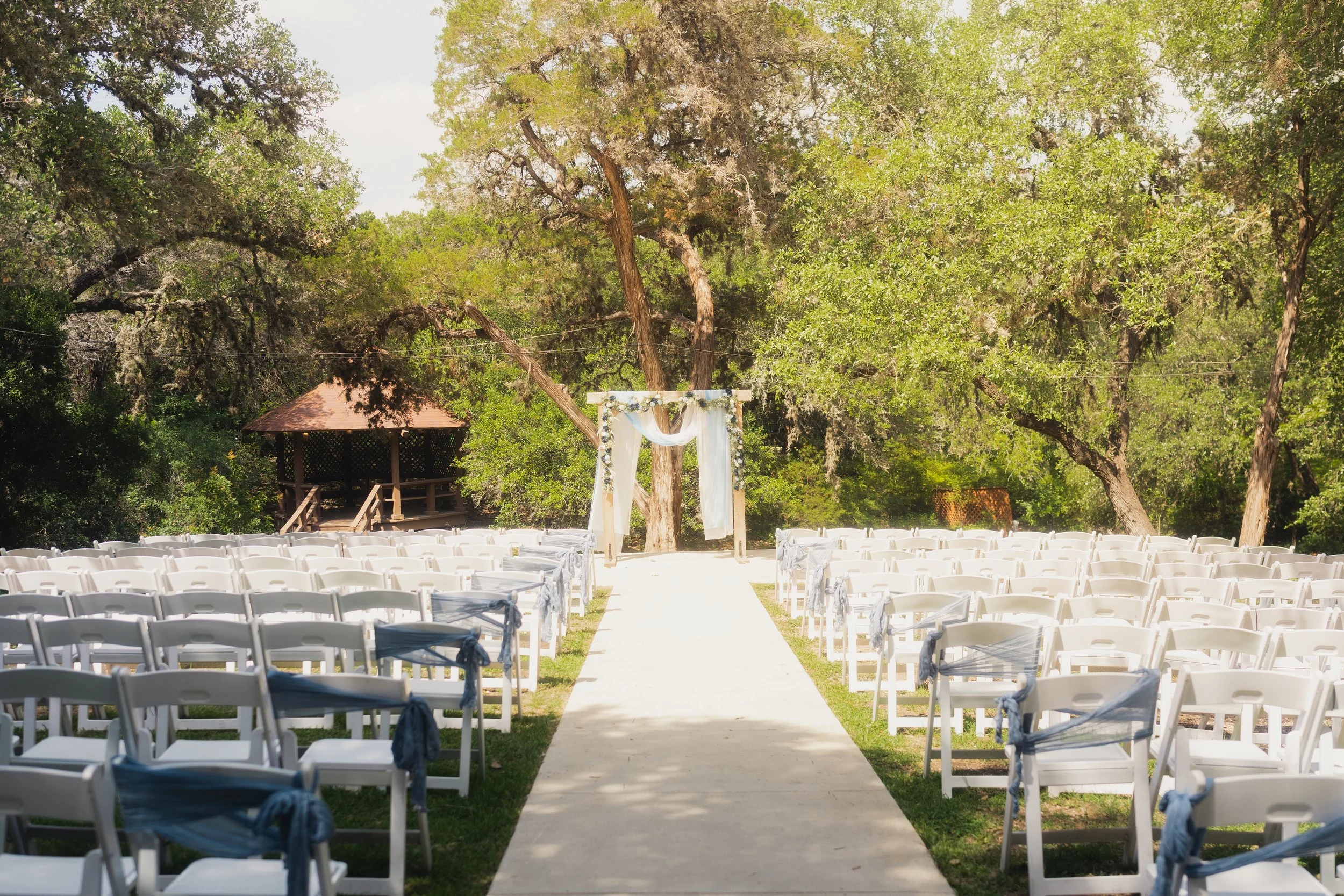 Outdoor wedding ceremony setup with white chairs tied with blue sashes, an arch decorated with white drapes and flowers, surrounded by trees and a wooden gazebo in the background.
