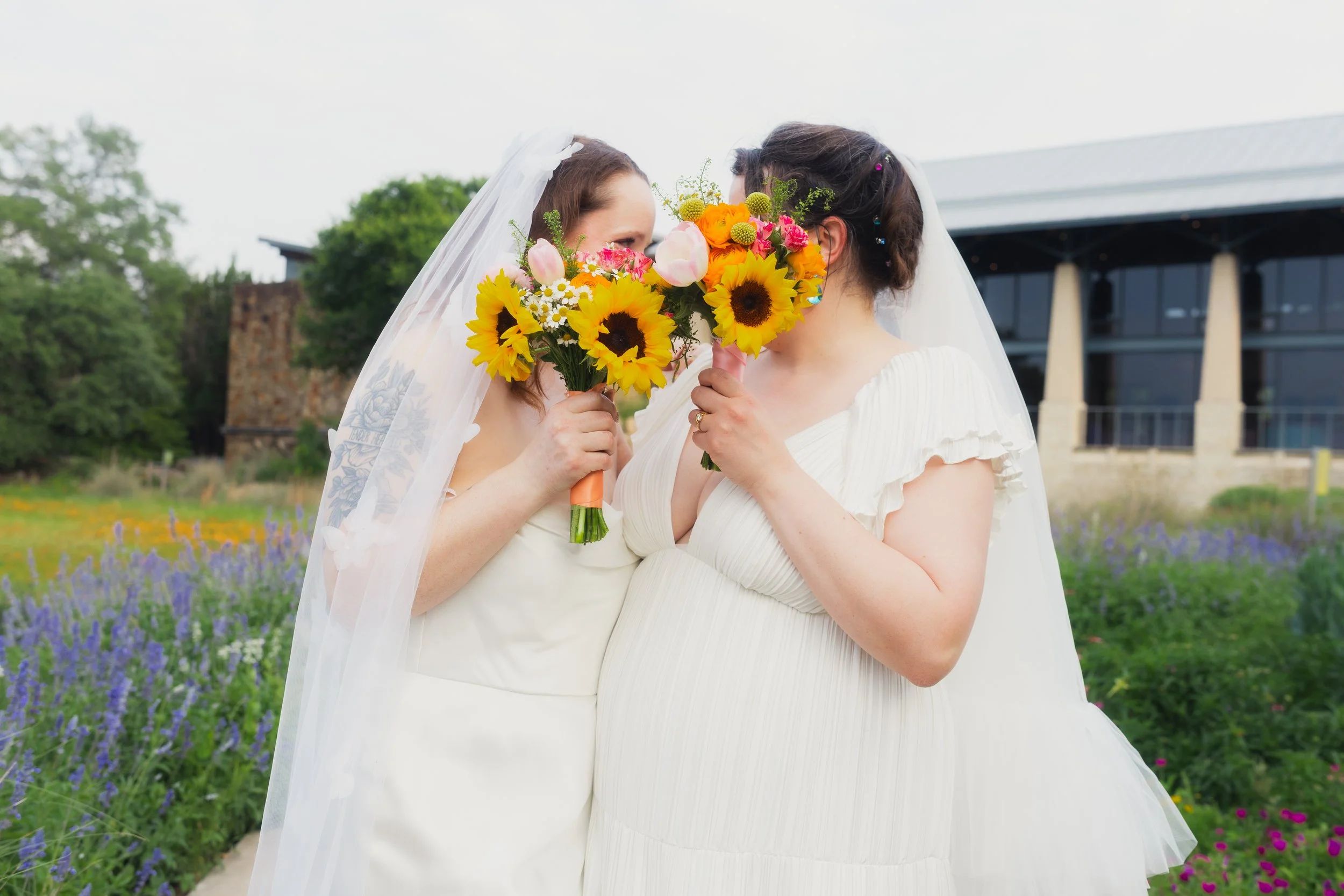 Two women in wedding dresses holding colorful bouquets, standing close and facing each other outdoors with a garden and modern building in the background.