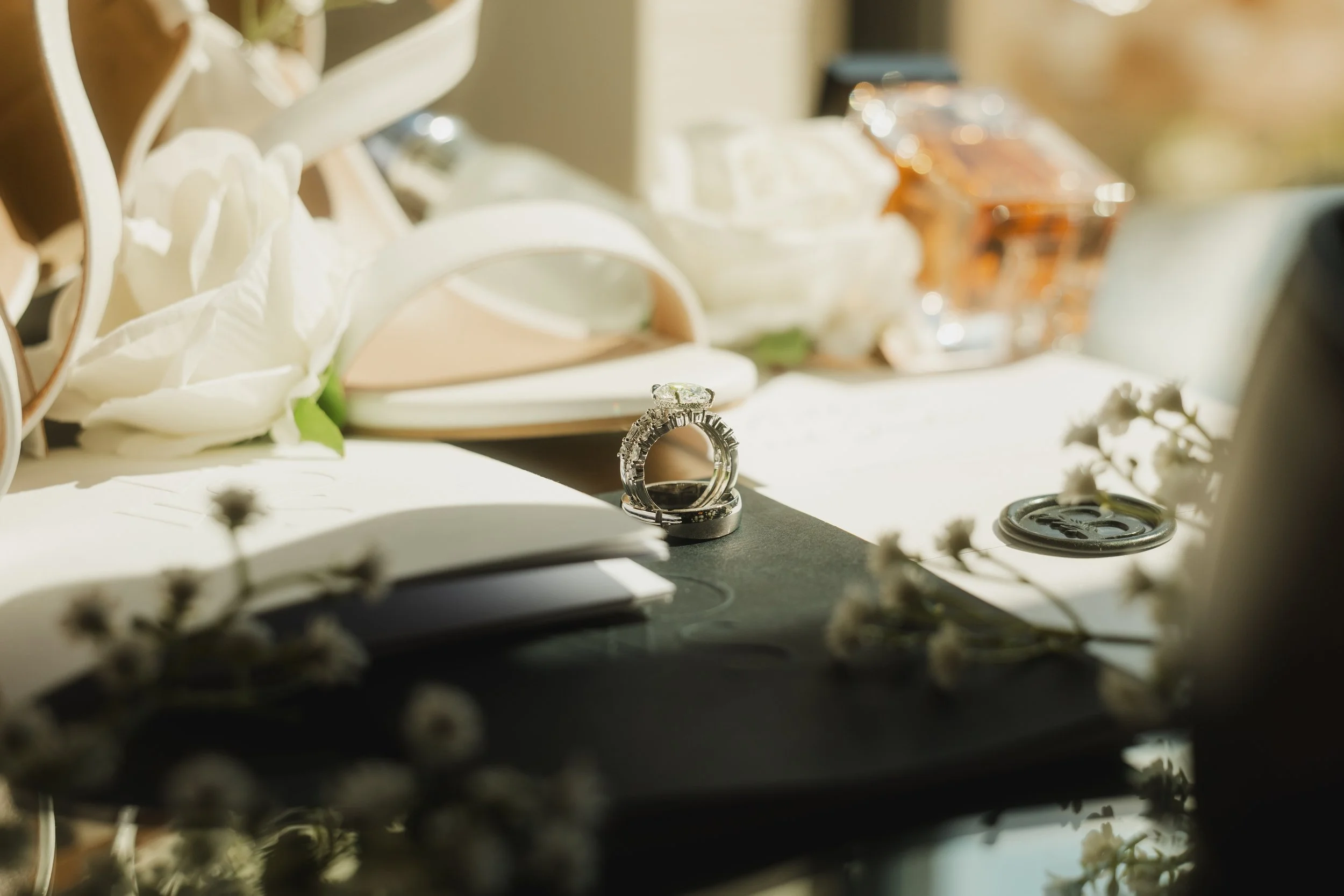 Close-up of wedding rings on a black surface with a blurred background of flowers, jewelry, and miscellaneous items.