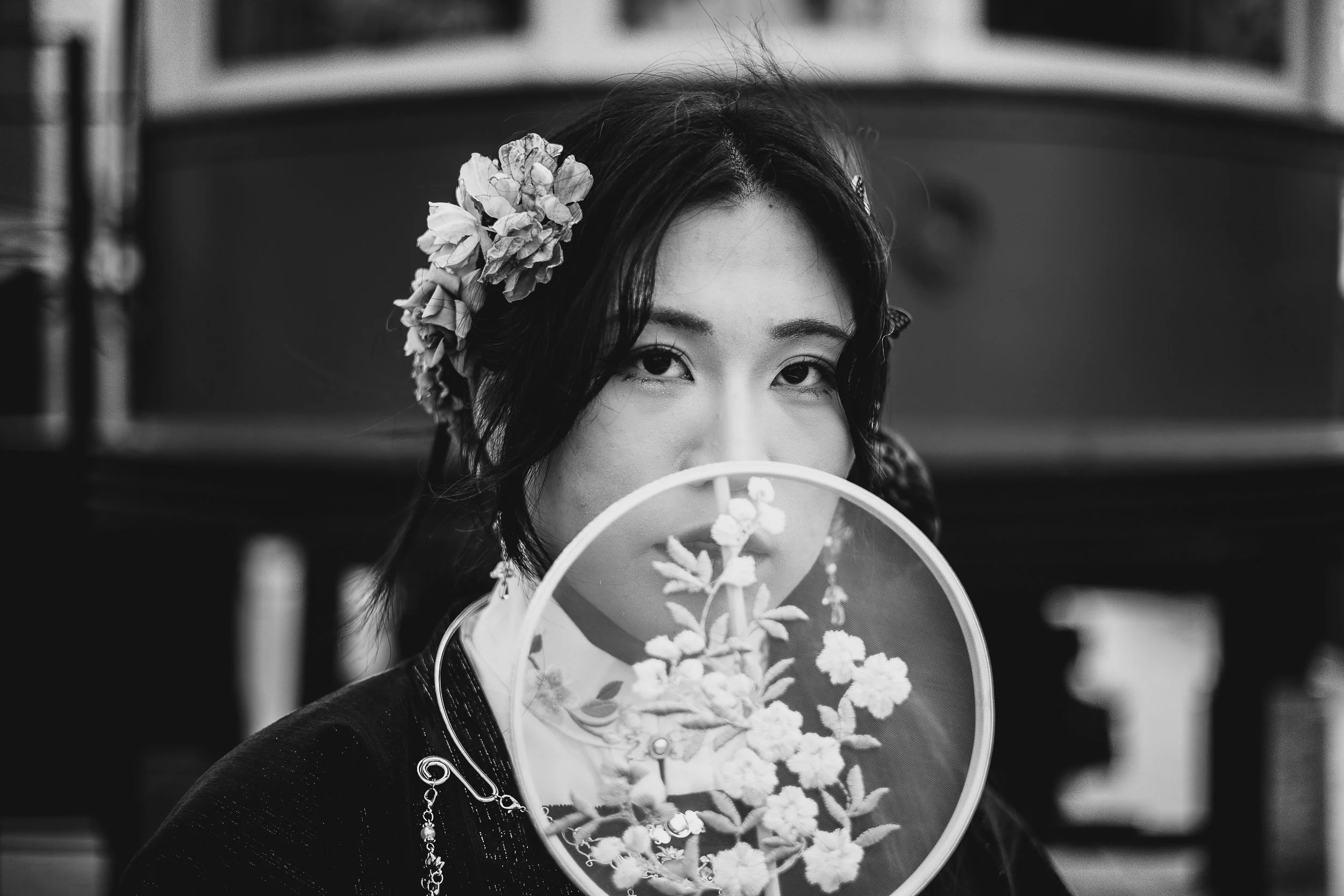 A woman with floral hair accessories holds a decorative fan in front of her face, partially obscuring her features. She looks directly at the camera with an intense expression.