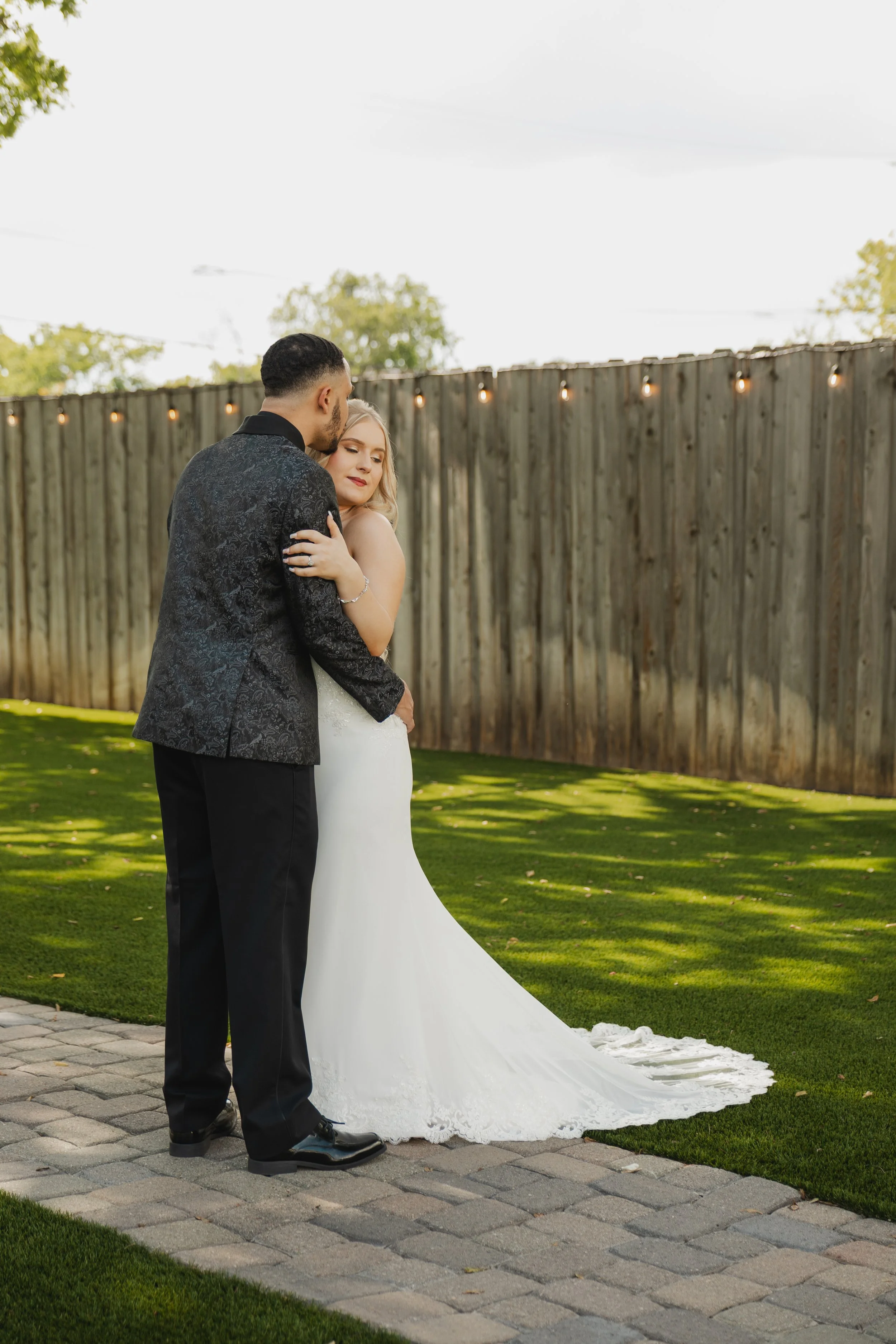 A bride and groom hugging outdoors on a wedding day, with the bride in a white gown and the groom in a black tuxedo, on a paved walkway near a wooden fence with string lights.