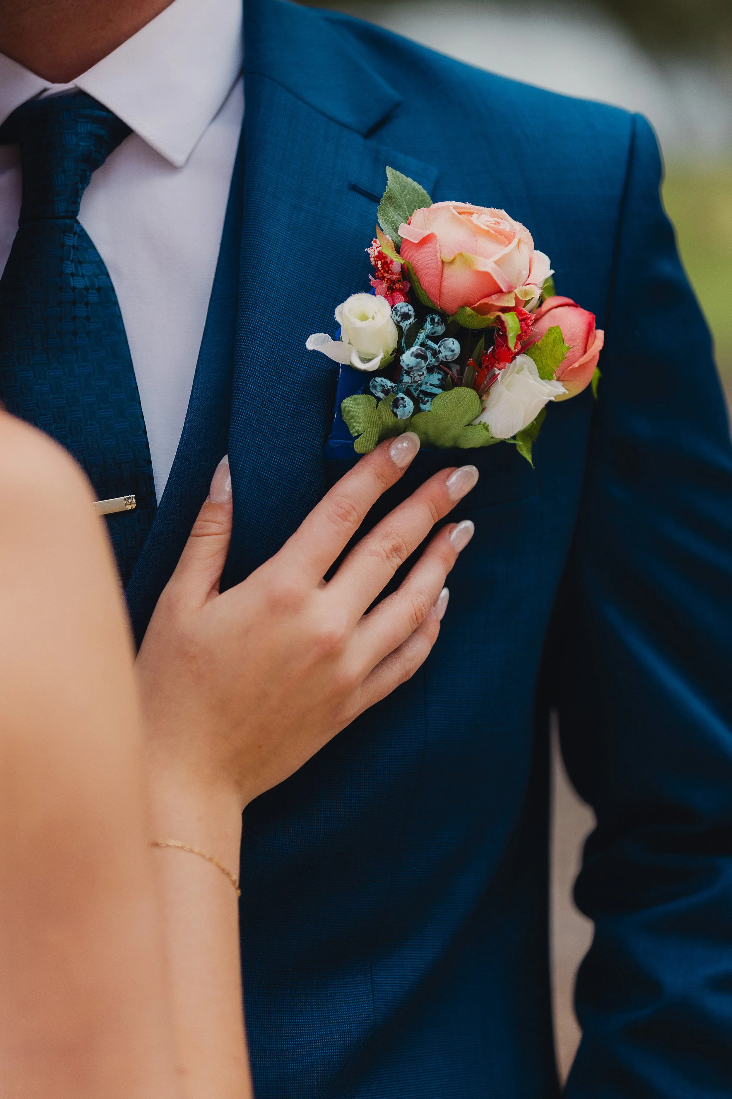 Person wearing a blue suit with a boutonniere of roses, berries, and greenery, while a hand with painted nails and a gold bracelet touches the suit.