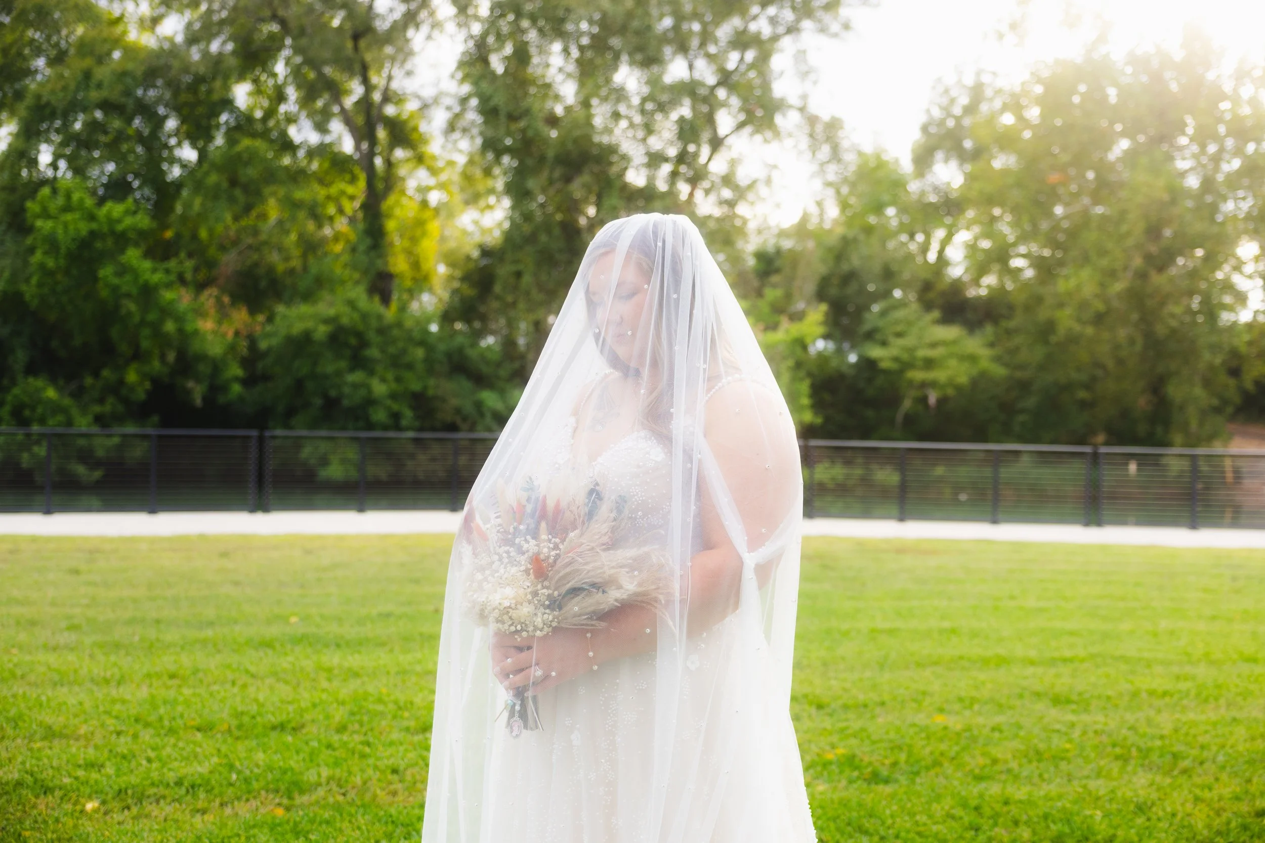 A bride stands in a grassy field, wearing a white wedding dress and veil, holding a bouquet, with trees and sunlight in the background.