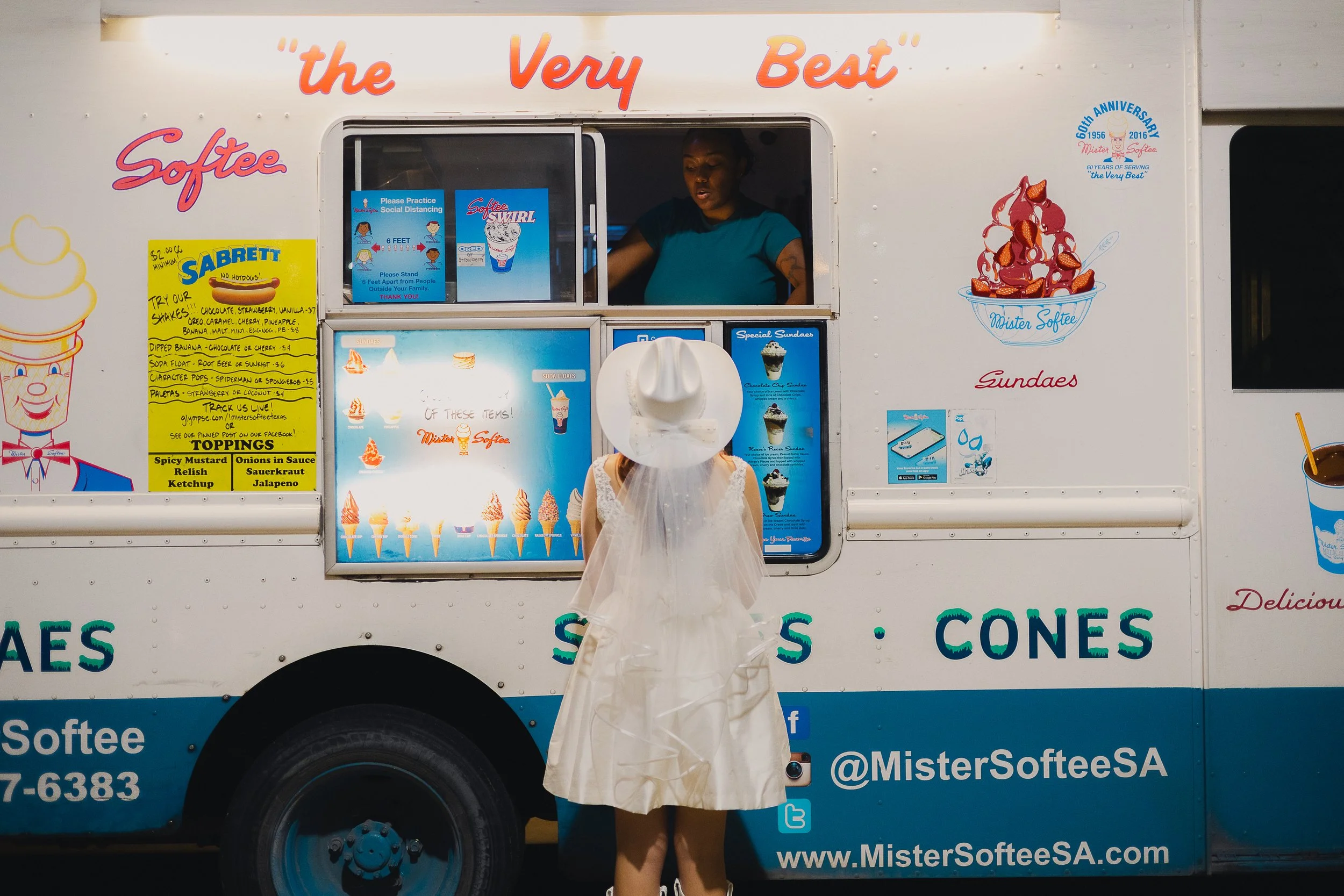 A woman in a white dress and hat stands in front of a soft serve ice cream truck, looking inside at the vendor. The truck has colorful signage advertising ice cream and toppings, with a yellow menu on the left side, and cartoon illustrations of ice c