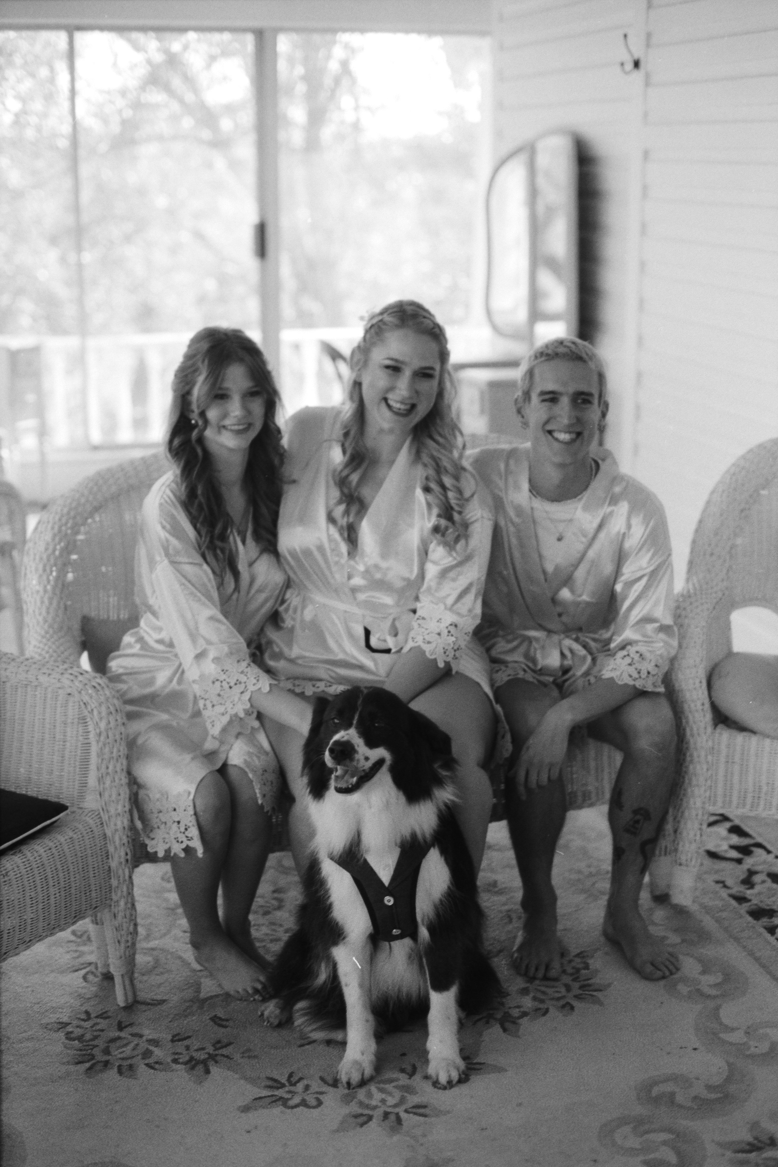 Three women in satin robes sitting on wicker furniture with a black and white dog in front of them inside a bright room with large windows.