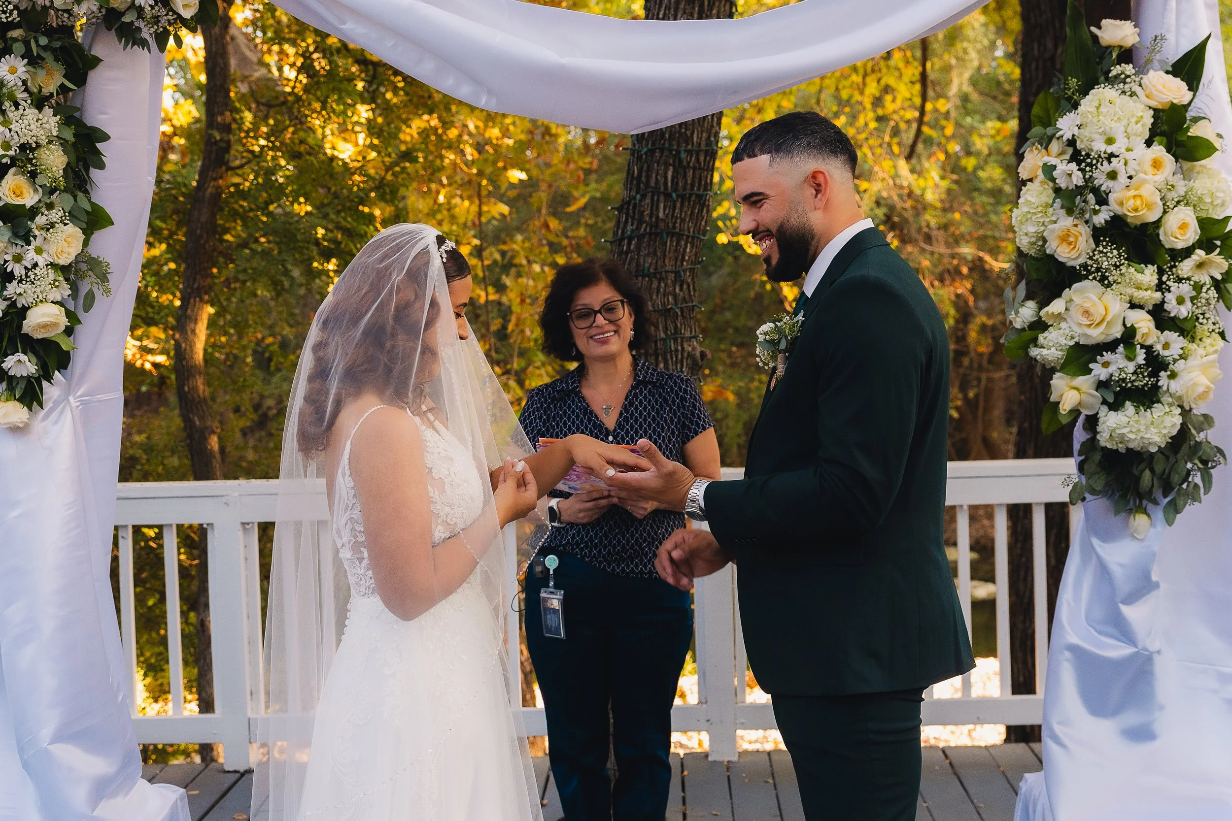 A couple getting married outdoors under a decorated arch with white fabric and floral arrangements during sunset. The bride is wearing a white lace wedding dress and a veil, and the groom is dressed in a black suit with a boutonnière. A woman, possib
