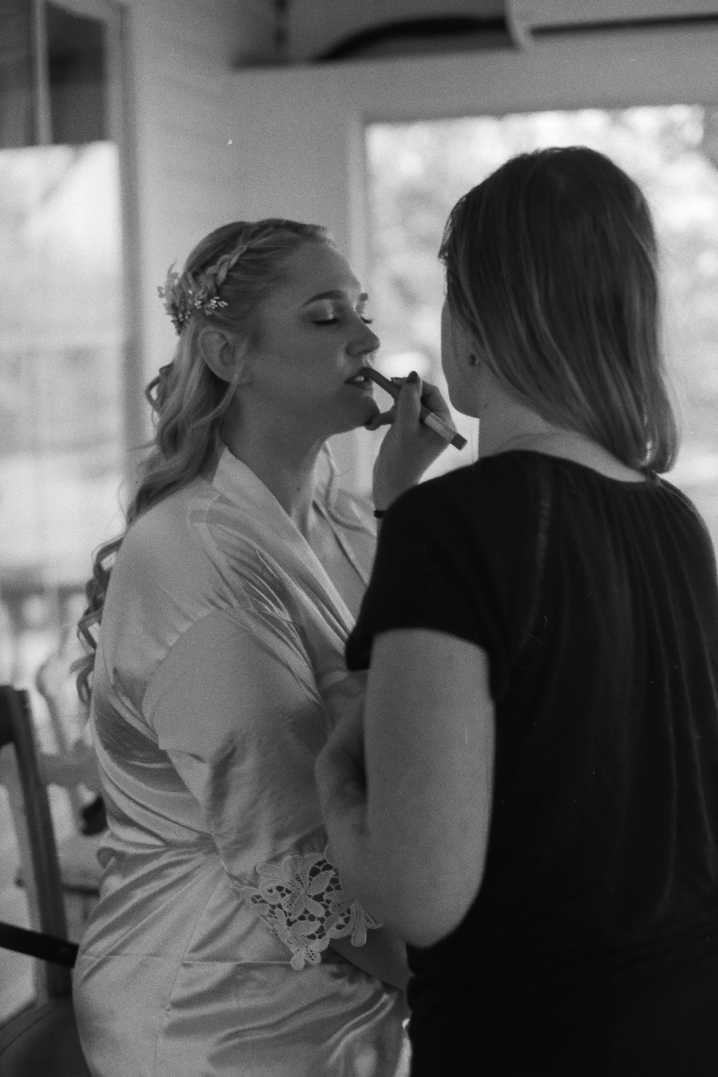 A woman with light hair styled with braids and a flower hair accessory is having her makeup applied by a makeup artist with dark hair. The makeup artist is using a brush on her lips. The scene appears to be in a well-lit indoor setting.