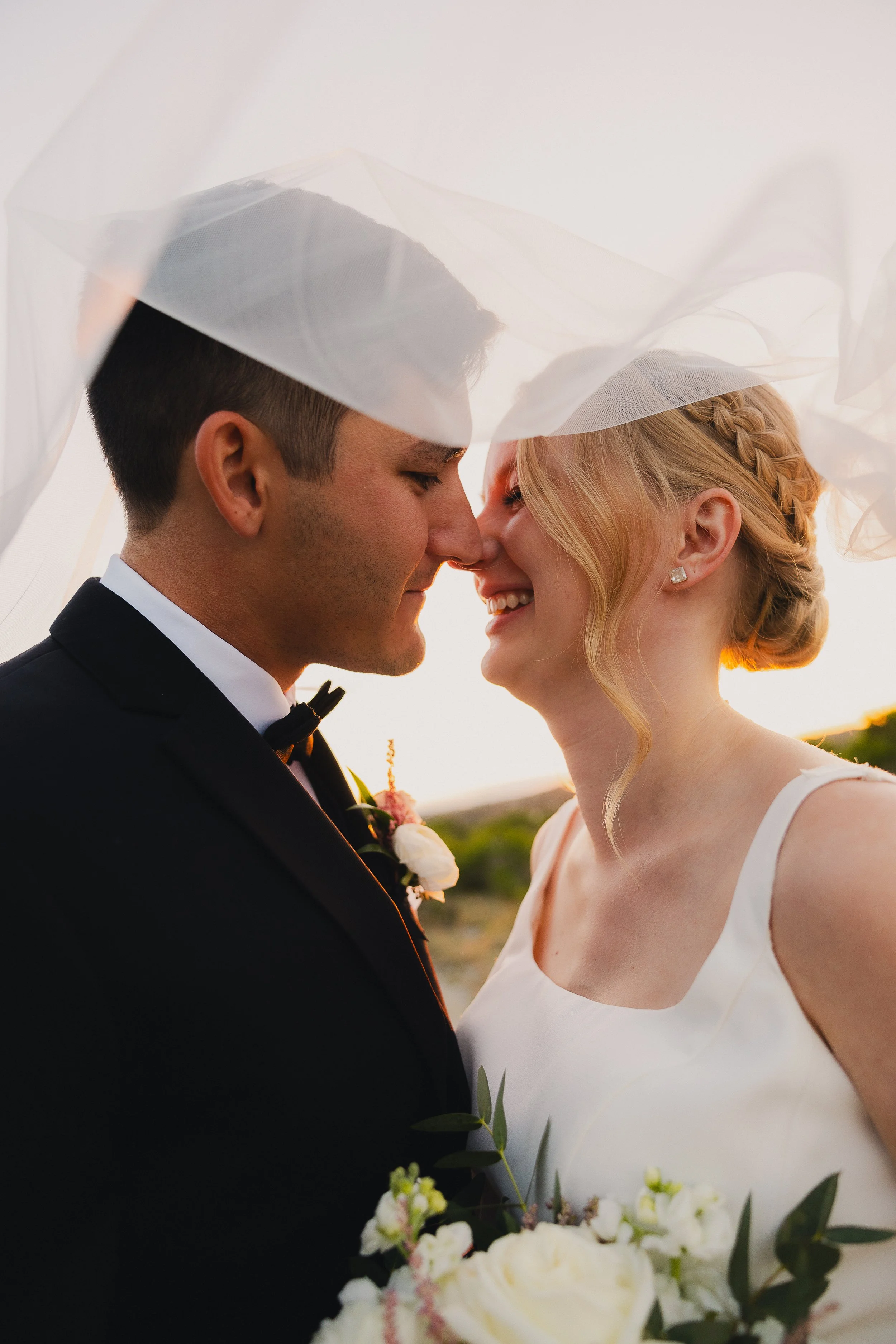 A bride and groom are close together, smiling and nearly touching noses, under a sheer veil at sunset, with the bride holding a bouquet of white flowers.