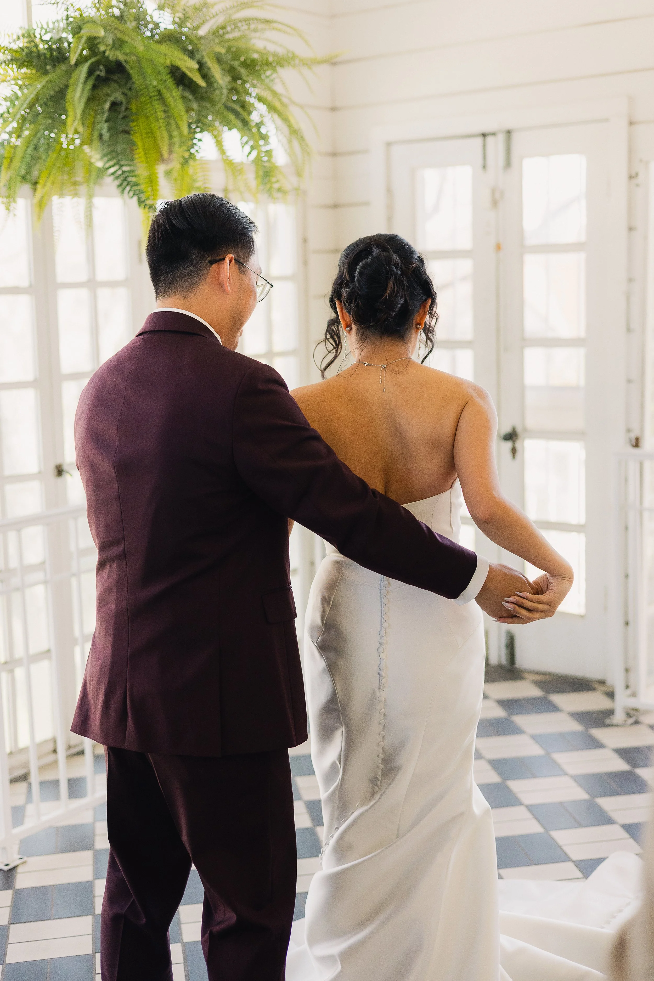 A couple is dancing, with the man in a dark suit and the woman in a strapless white wedding dress, inside a bright room with large windows and a checkered floor.
