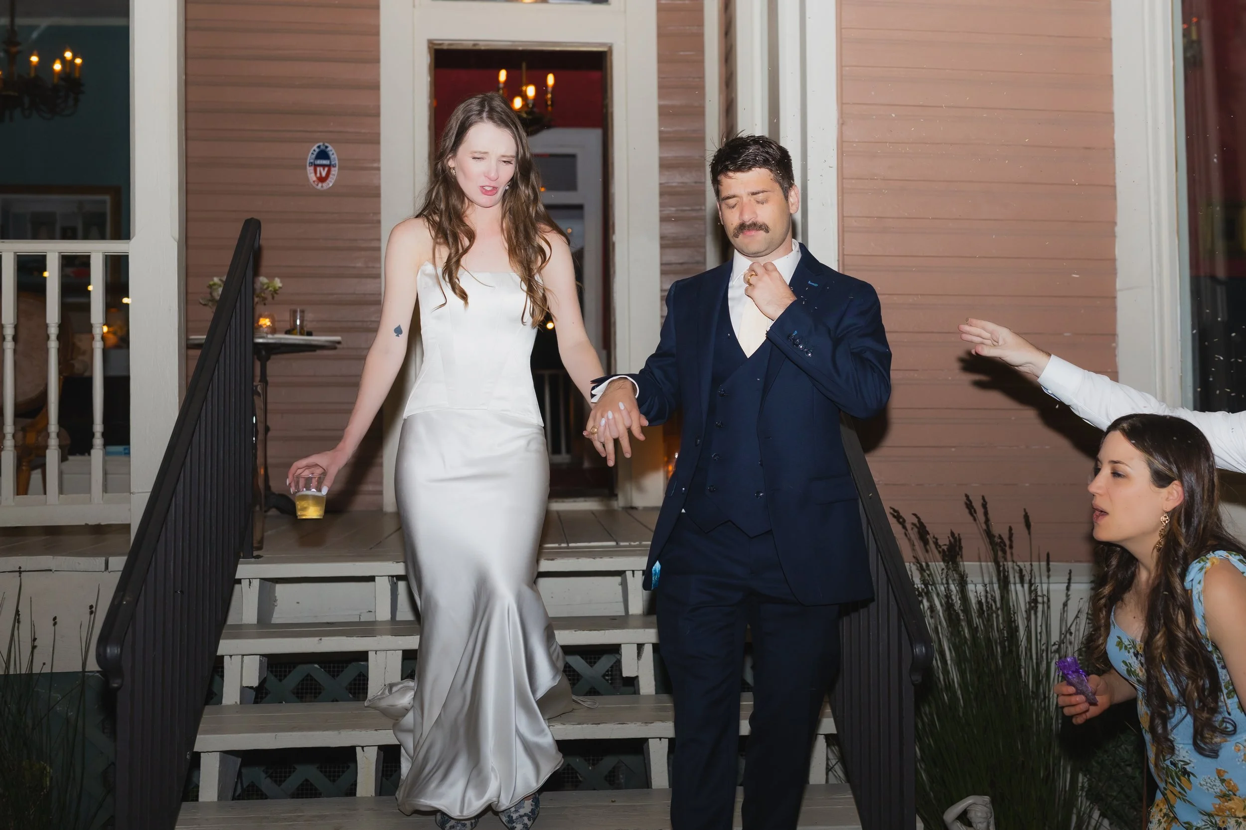 A woman in a white dress descending stairs holding a drink, accompanied by a man in a navy suit and tie, with a woman in a floral dress reaching out nearby at a night event.