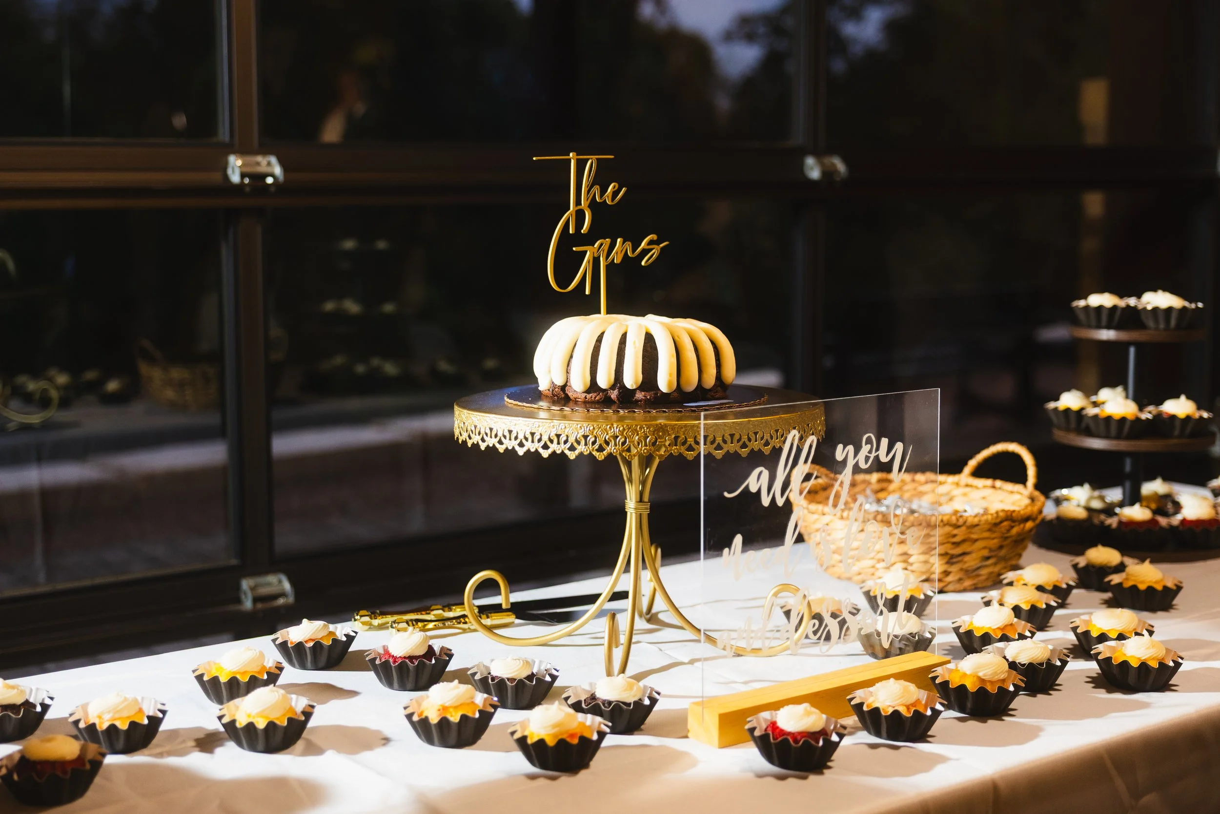 Decorative dessert table featuring a round cake with white and dark chocolate drizzles, topped with a gold cake topper that says 'The Gans.' The table is surrounded by numerous mini cupcakes with white frosting in black and gold wrappers, a large wov