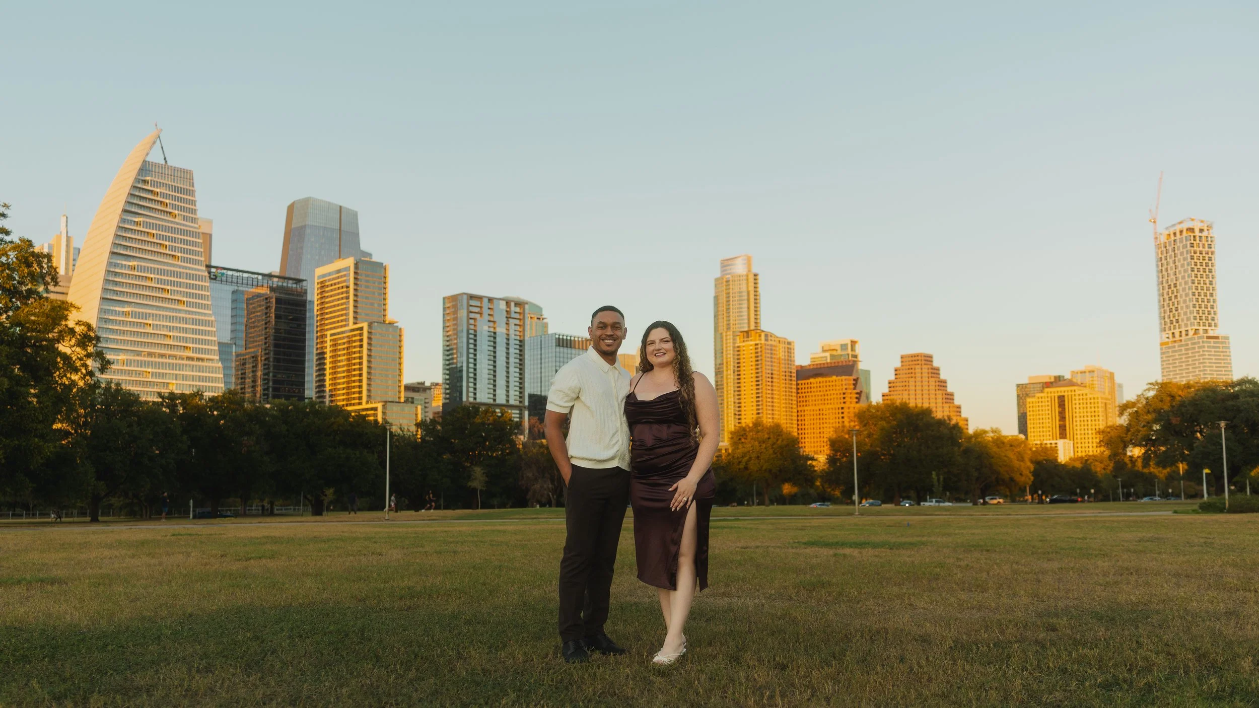 A smiling couple standing together outdoors in a park with a city skyline in the background during sunset. The man is wearing a white sweater and the woman is in a dark purple dress with a slit.