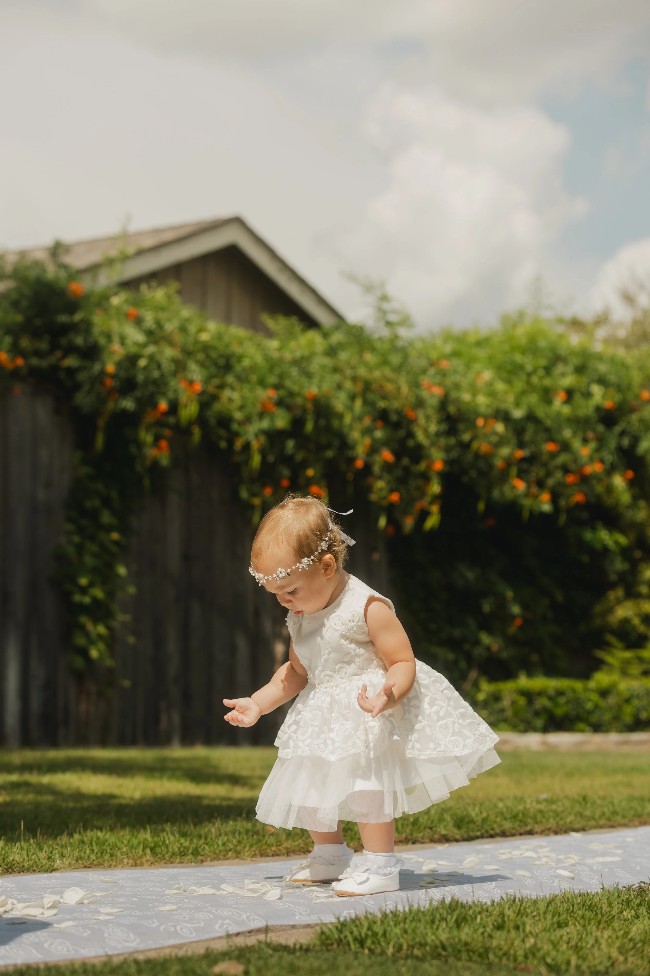A young girl in a white dress and floral headband bows her head while walking on a white patterned cloth outdoors on a grassy area on a sunny day