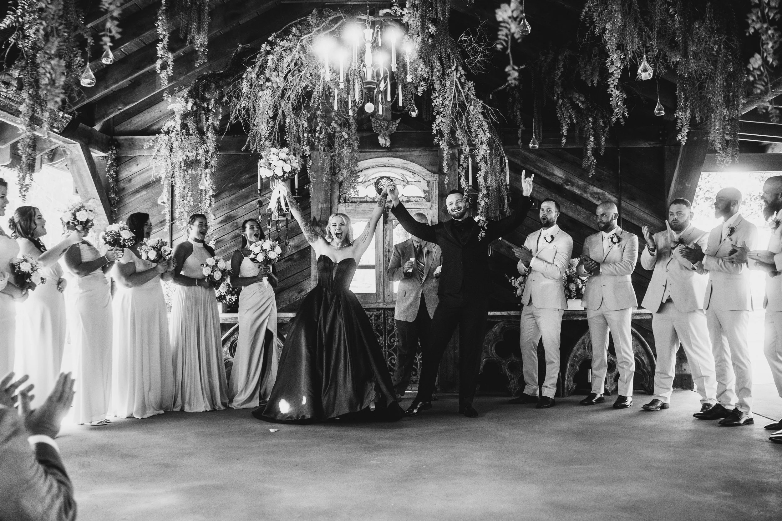 Black and white photo of a wedding celebration in a rustic hall with wood-paneled walls and a chandelier. The bride and groom are in the center, holding hands and smiling. The bride wears a strapless gown and the groom a dark suit. Bridesmaids in mat