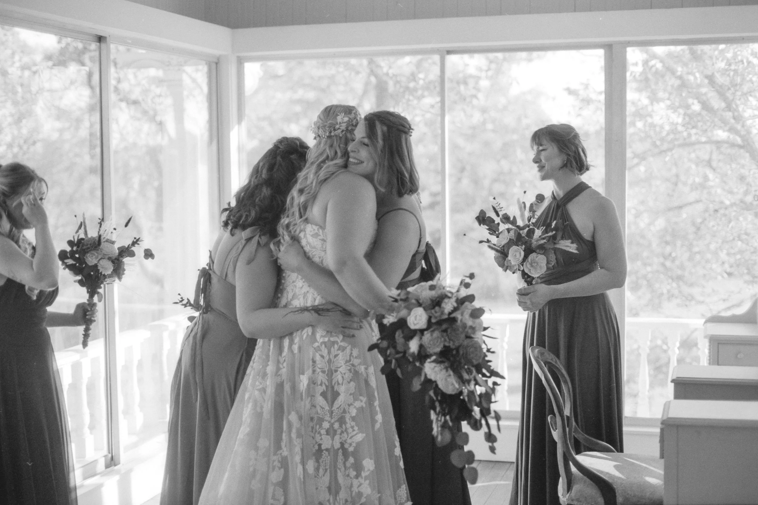 A group of women, including one bride in a wedding dress, hugging among other bridesmaids holding bouquets, with another bridesmaid holding a bouquet and smiling, in a bright room with large windows and trees outside.
