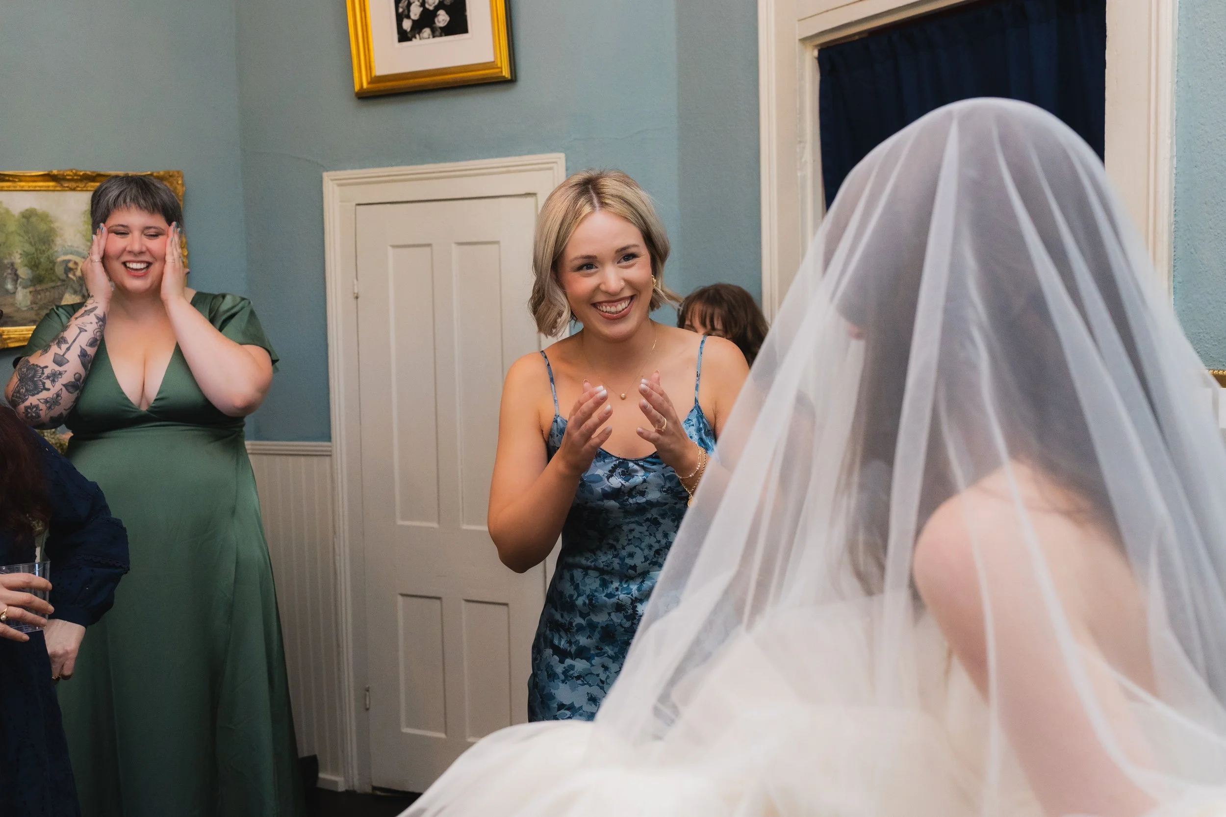 A bride in a wedding gown with a veil sitting during a gathering with three women expressing joy and surprise, one woman in a blue dress smiling and another in a green dress with tattoos on her arms covering her ears.