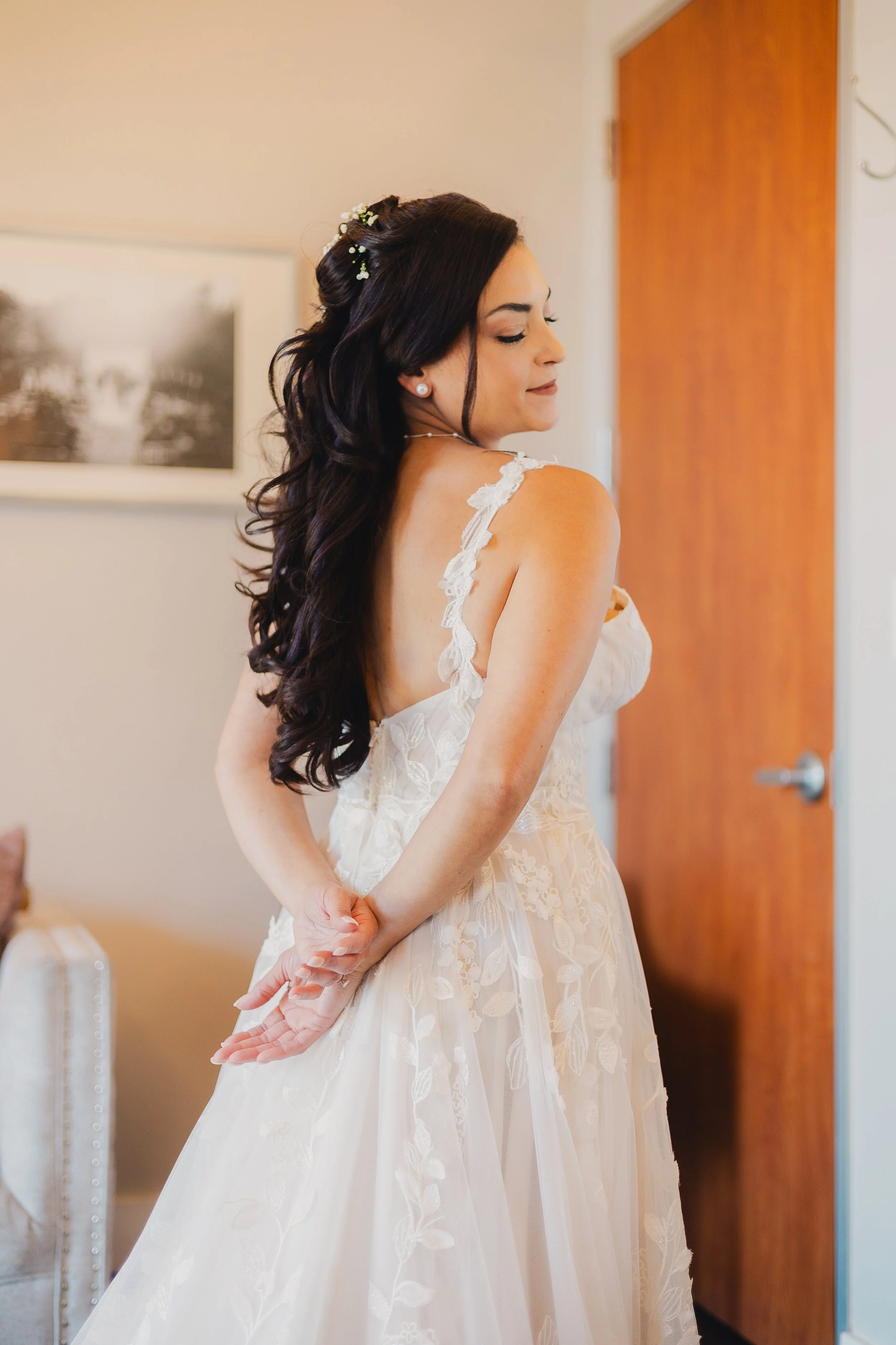 A woman with long dark hair, dressed in a white lace wedding gown, stands with her hands clasped behind her back in a softly lit room, appearing to prepare for her wedding.