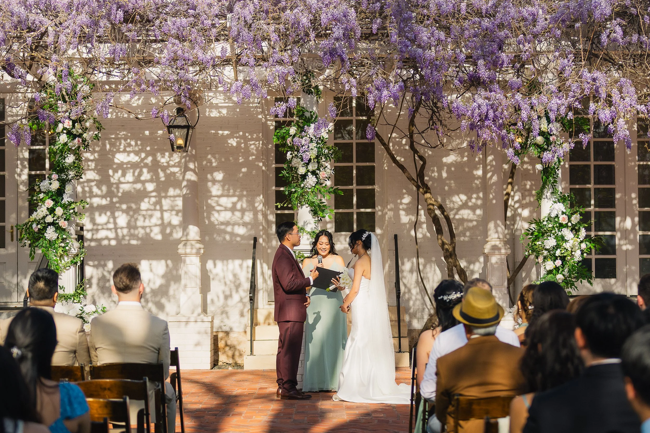 A wedding ceremony taking place outdoors under a decorated arch with purple wisteria and white flowers. The bride and groom stand facing each other, with the officiant in the middle. Guests are seated, witnessing the ceremony.