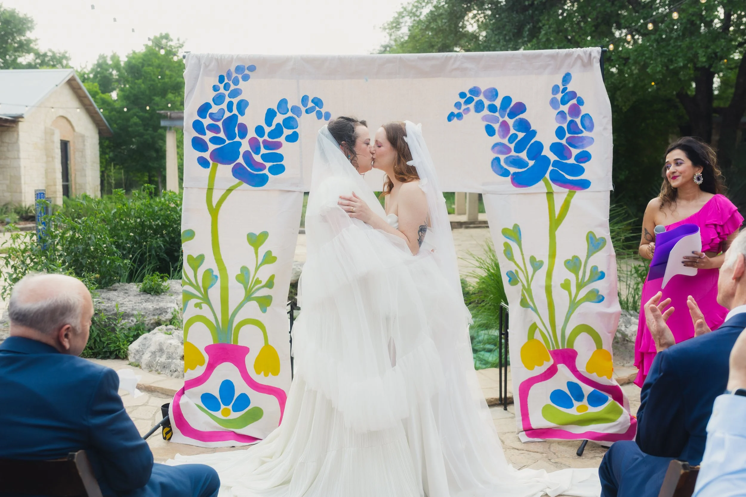 Two brides kiss during a wedding ceremony outdoors, with a painted floral backdrop, surrounded by seated guests and a woman in a pink dress holding a microphone.