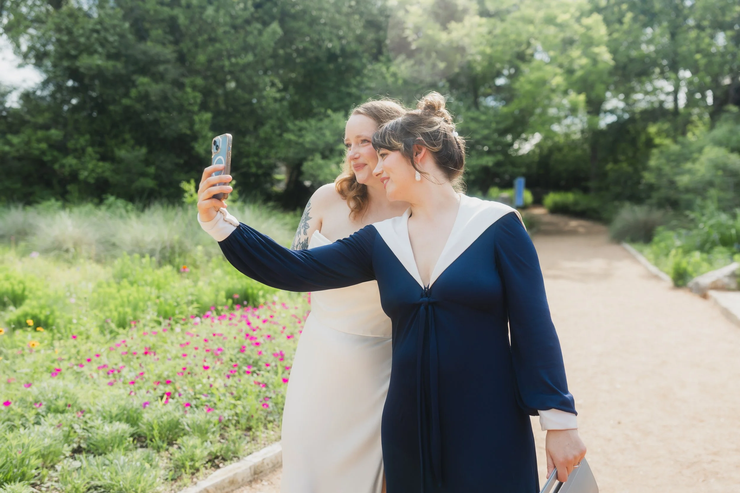 Two women taking a selfie outdoors in a lush, green park with flowers and trees.