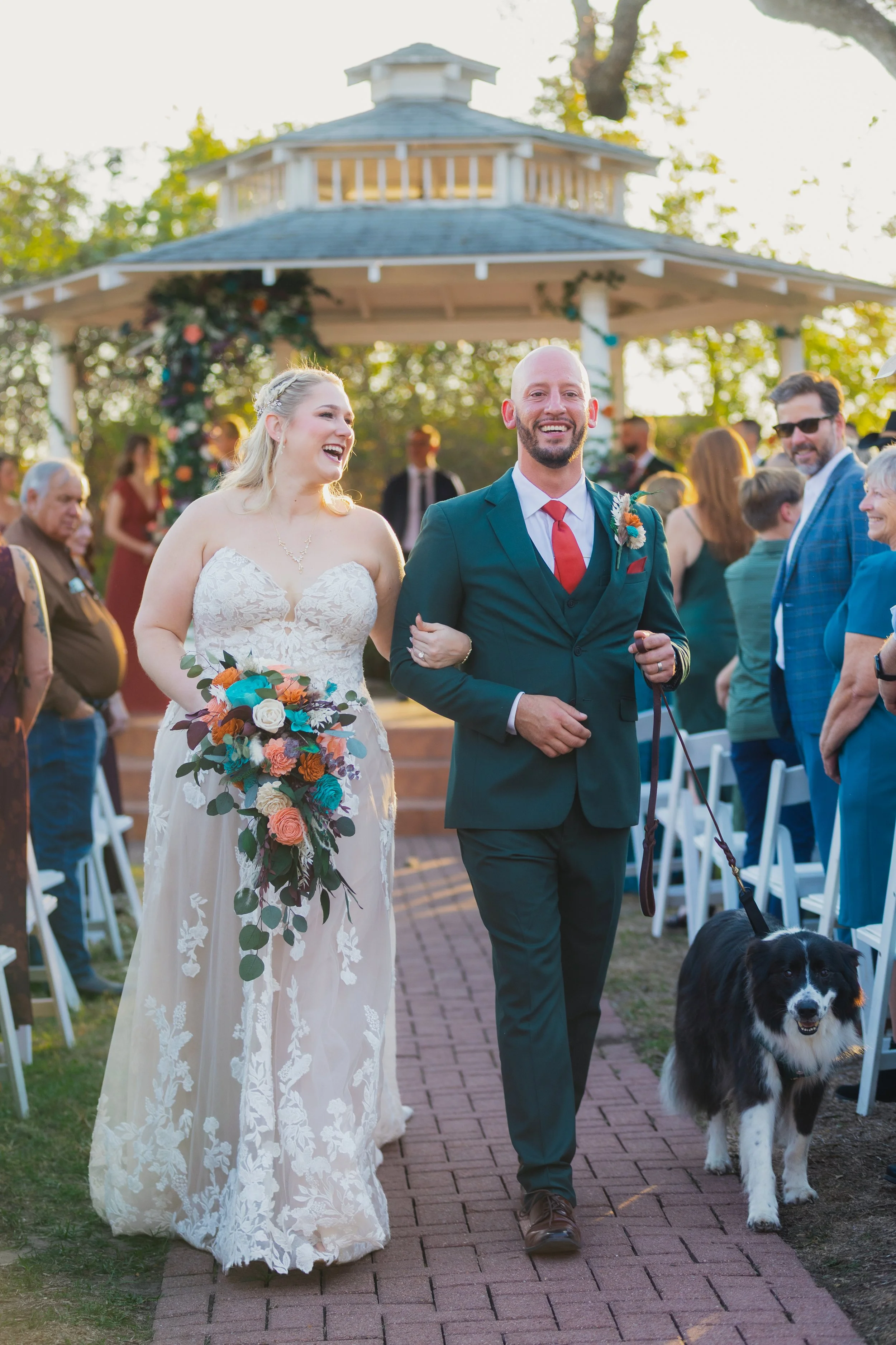A newlywed couple walking down an outdoor aisle, smiling and holding hands. The bride is in a lace wedding dress, holding a bouquet of colorful flowers. The groom is in a green suit with a red tie. Their dog is walking alongside them. Guests are stan