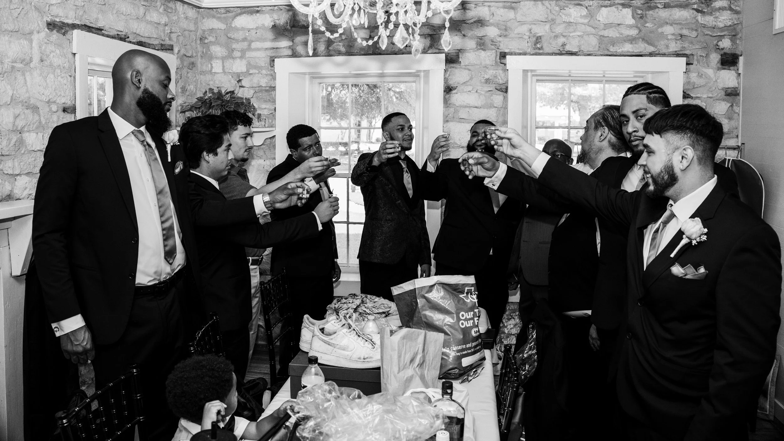 A group of men in suits raising glasses in a toast during a celebration in a room with stone walls, a window, and a chandelier.
