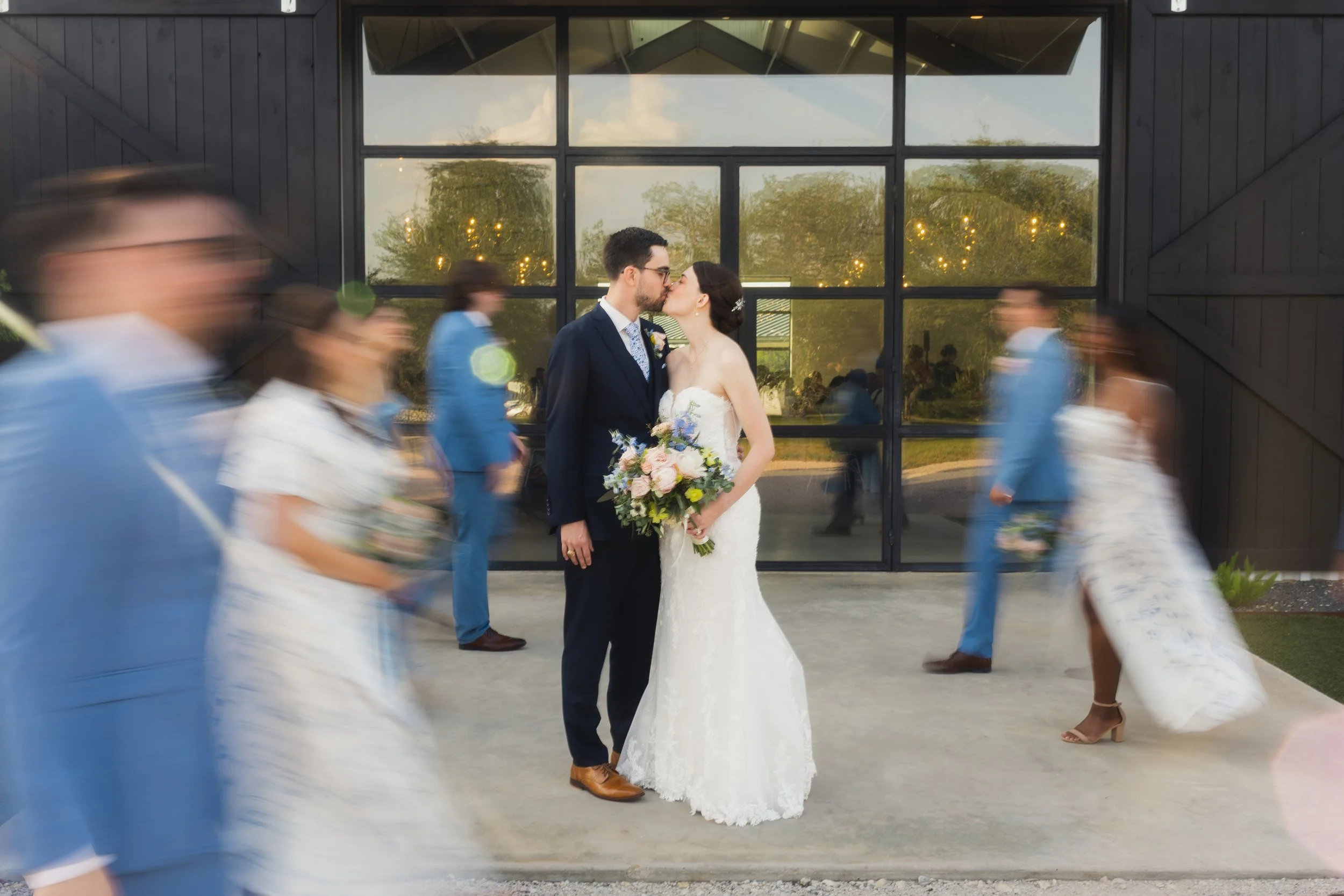 A couple in wedding attire stands facing each other and kissing, while wedding guests walk by in motion blur outside a dark modern building with large glass windows.