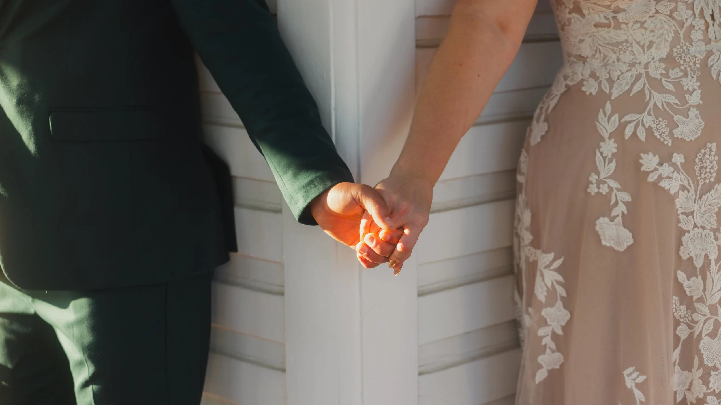 Close-up of a bride and groom holding hands during a wedding, standing against a white wooden wall.