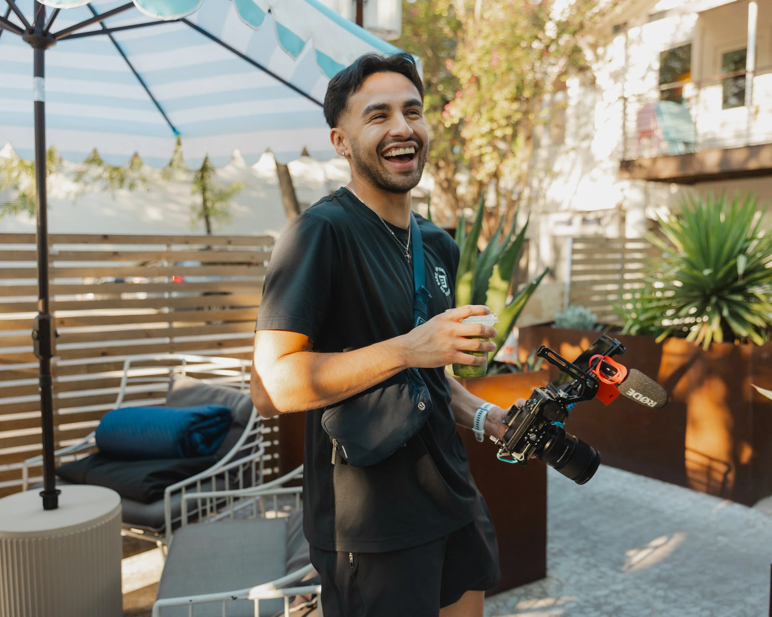 A man smiling and holding a camera and a drink, standing outdoors near plants and outdoor furniture.