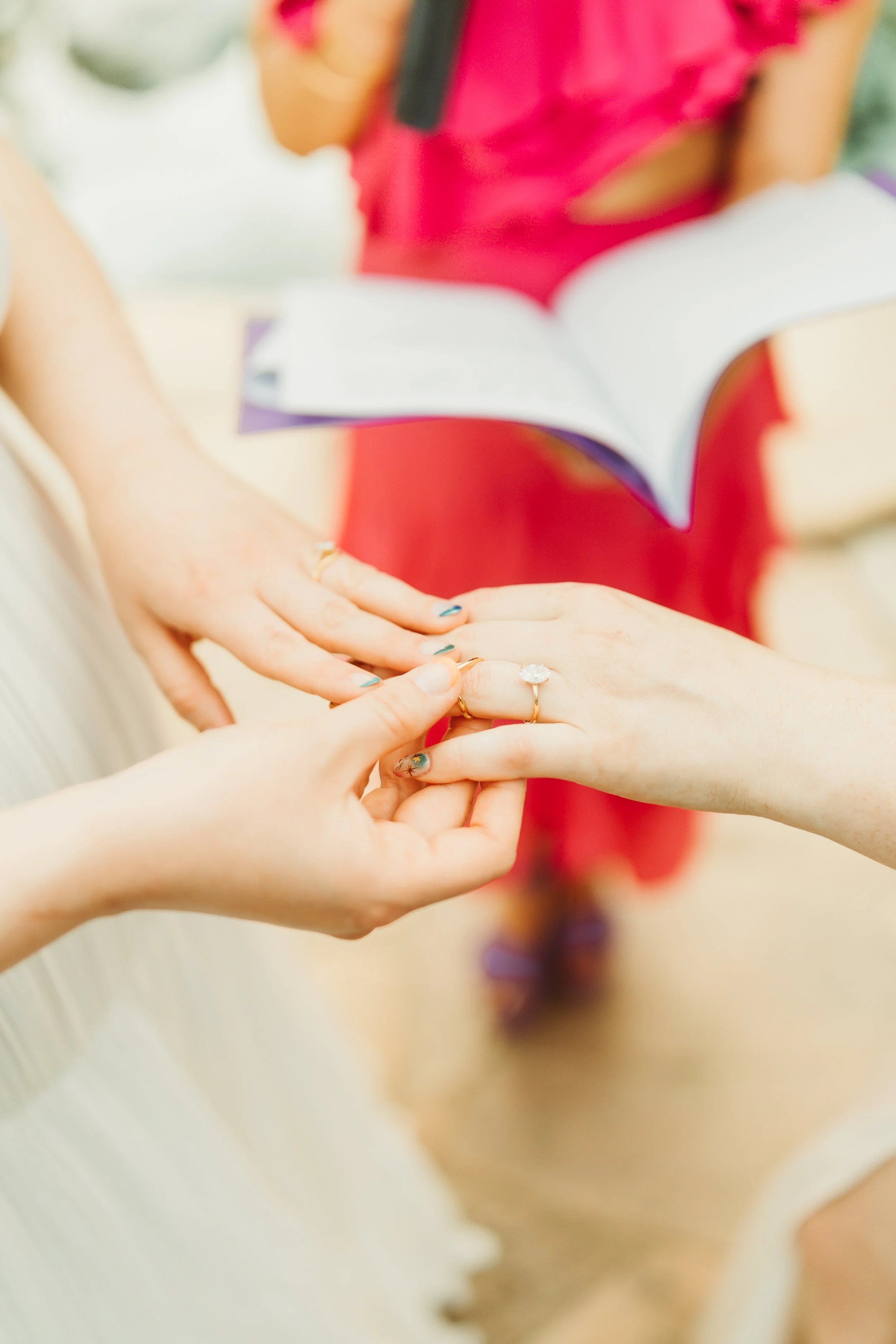 Three women exchanging rings, with a woman in a red dress holding an open book in the background.