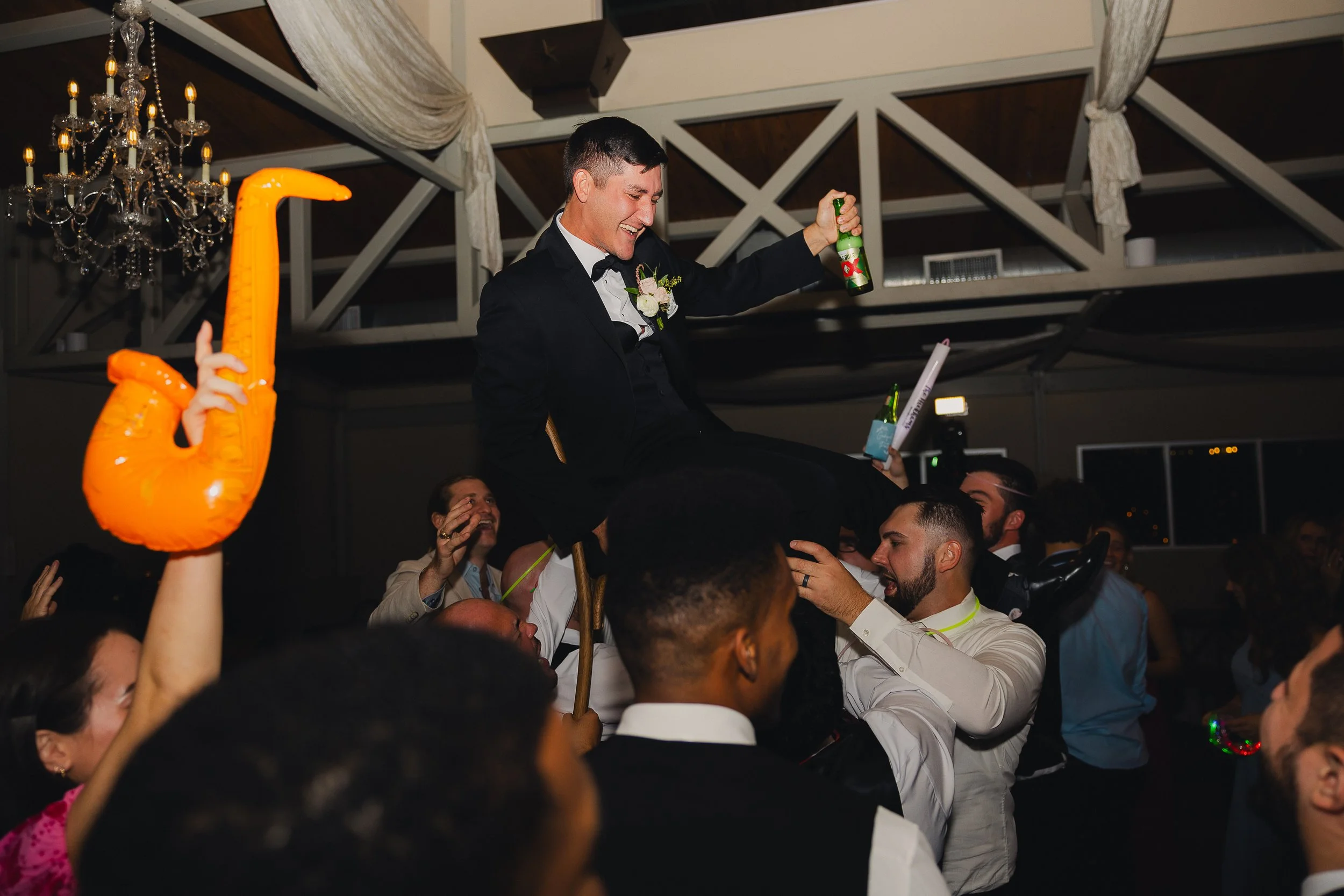 Man in a tuxedo sitting on a chair being lifted by friends during a celebration or wedding reception, surrounded by other guests cheering.