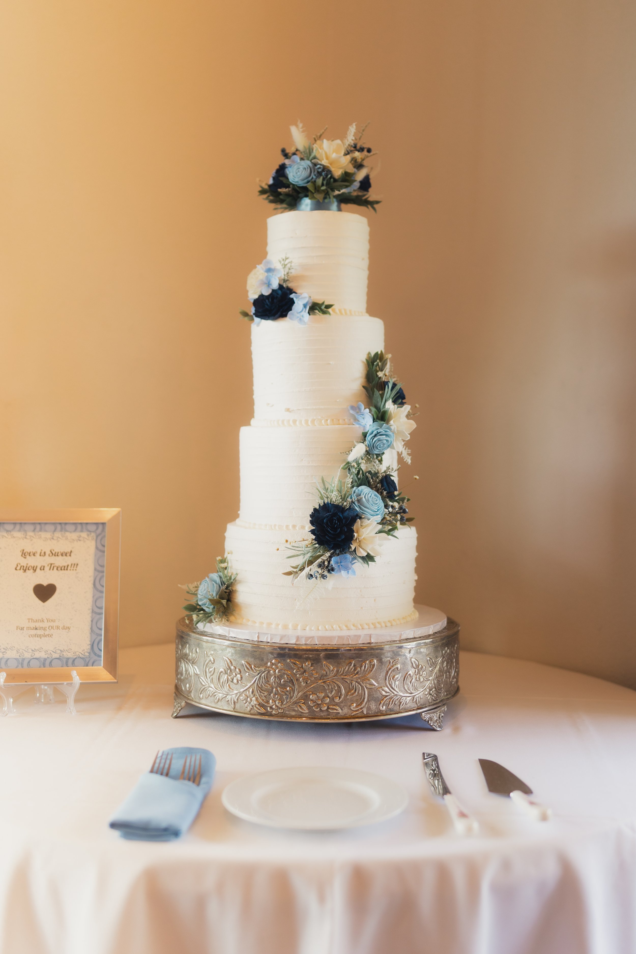 Elegant wedding cake with four tiers, decorated with blue and white flowers, on a silver stand at a reception table.
