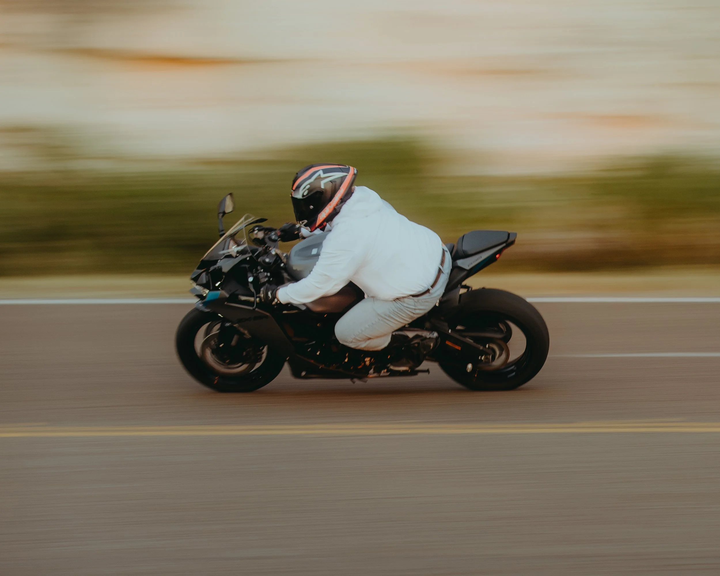Motorcycle rider dressed in a white jacket and beige pants, wearing a black helmet with orange accents, riding a black motorcycle on a road with motion blur background.