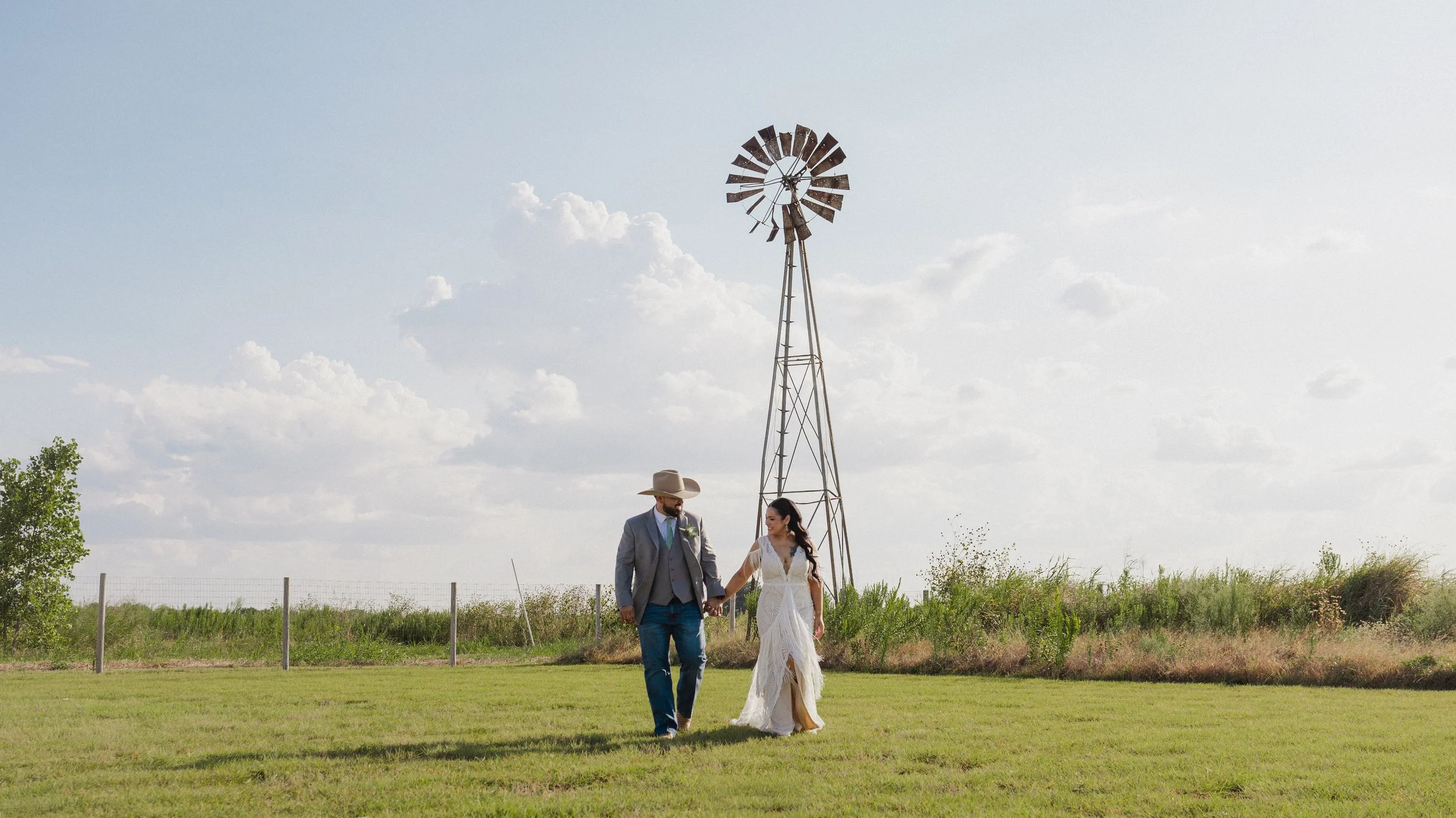A couple dressed in wedding attire walking hand in hand outdoors with a windmill in the background on a partly cloudy day.