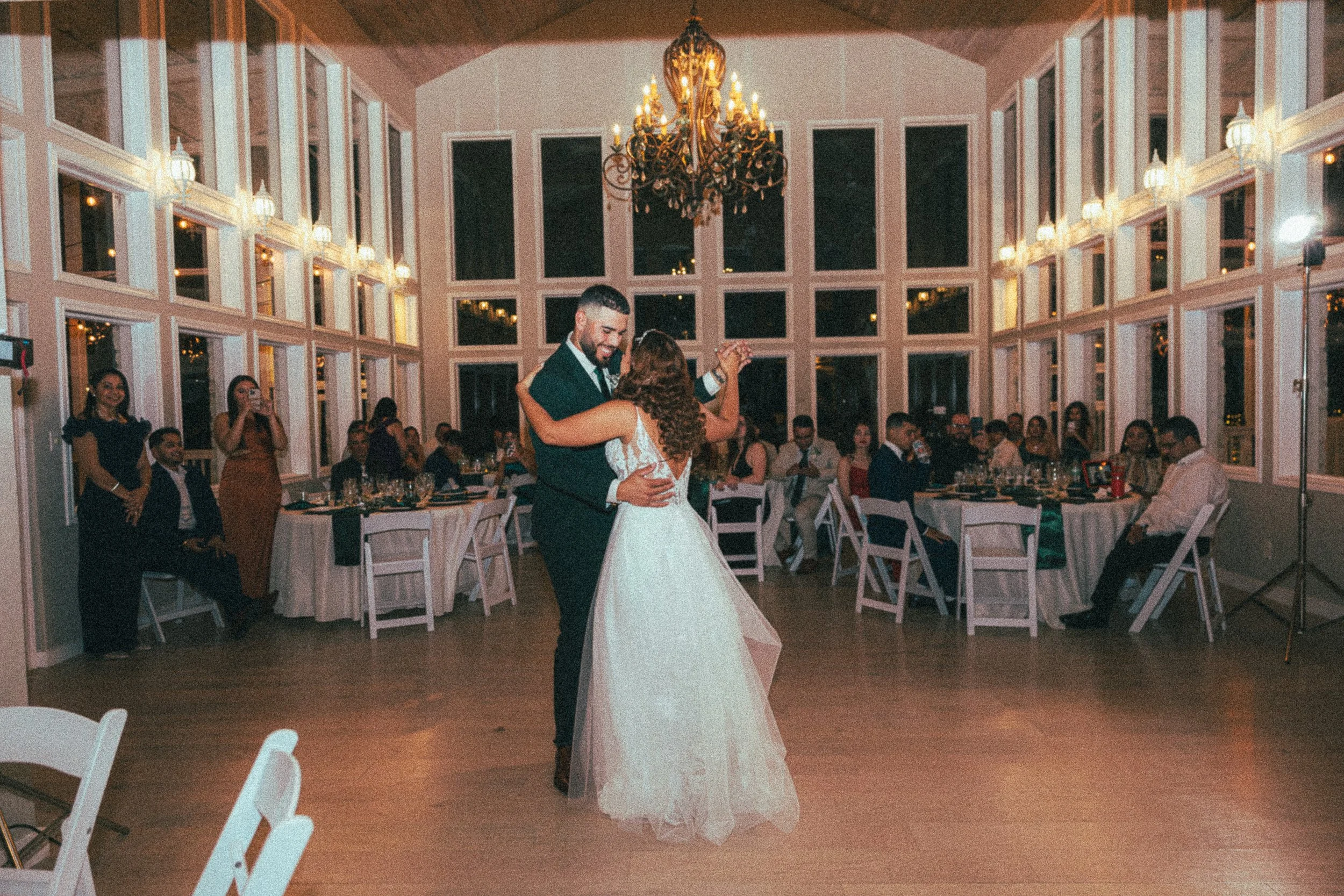 A bride and groom dance at their wedding reception in a bright, elegant room with a chandelier and large windows. Guests are seated at tables, watching and enjoying the moment.