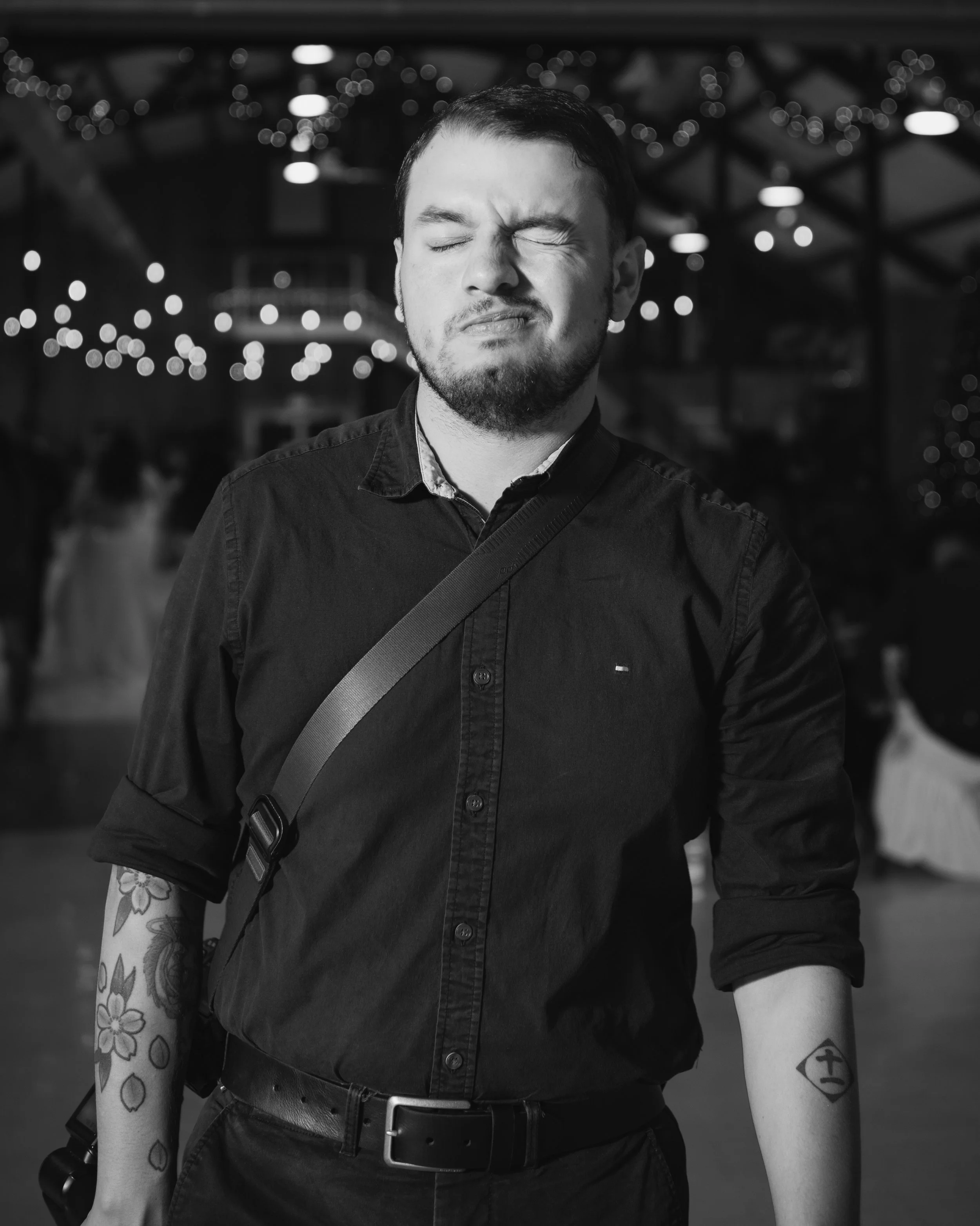 A man with closed eyes and a facial expression of discomfort or pain, wearing a black shirt with rolled-up sleeves, standing in an indoor setting with blurred lights in the background.