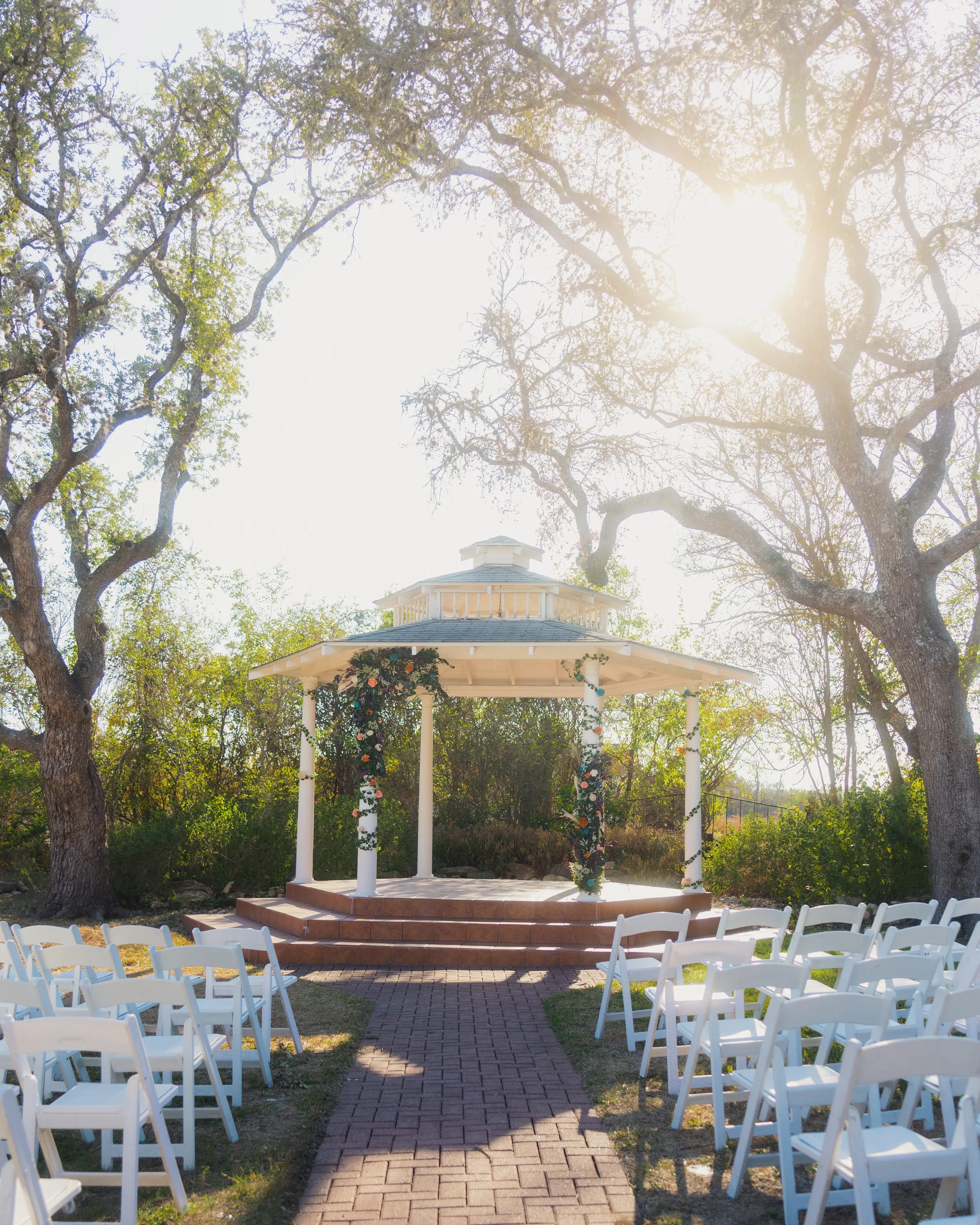 An outdoor wedding setup with a gazebo decorated with flowers and greenery, surrounded by white chairs, under trees with sunlight filtering through.