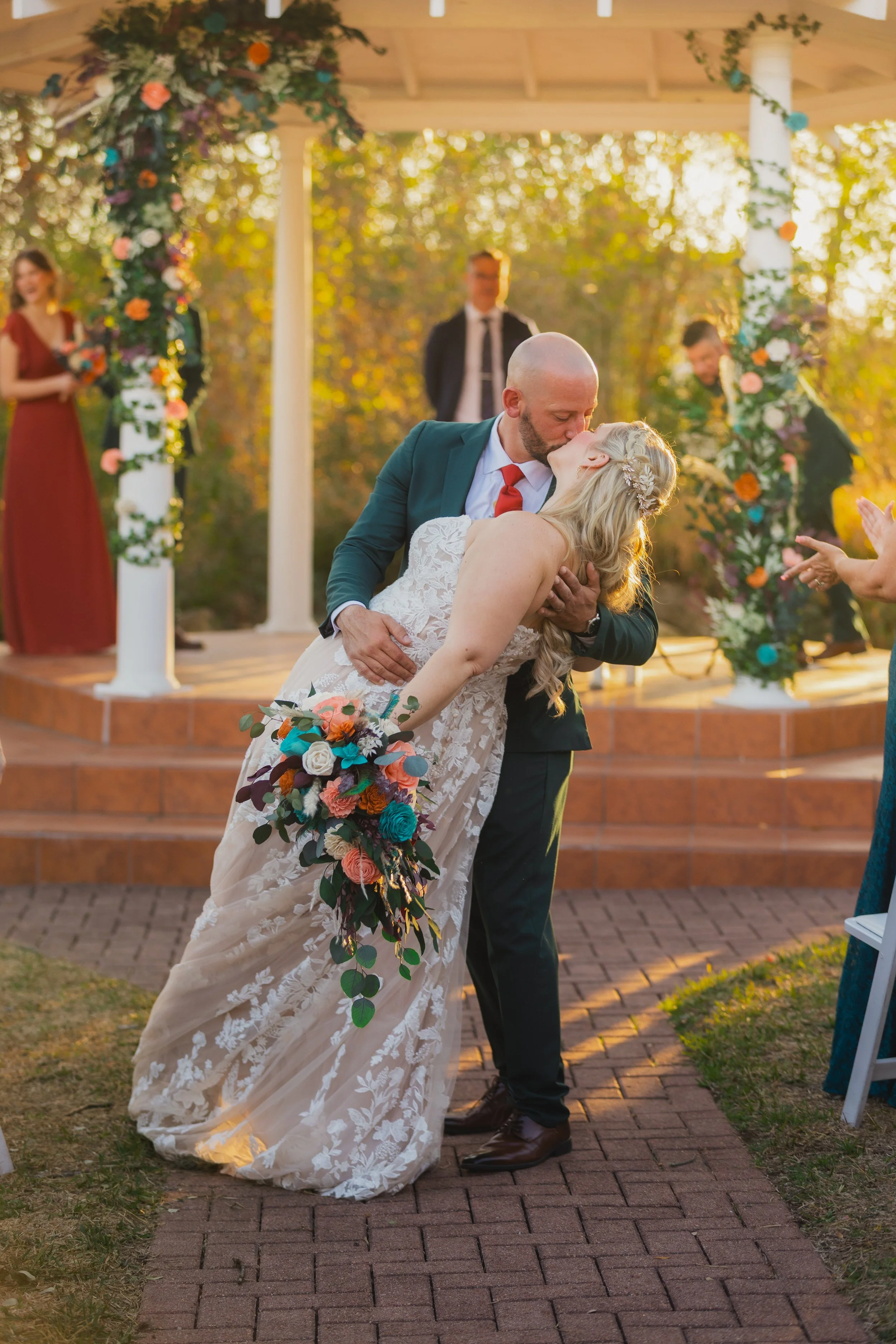 A couple kissing during their outdoor wedding ceremony, with a wedding arch decorated with colorful flowers in the background and bridesmaids standing nearby.