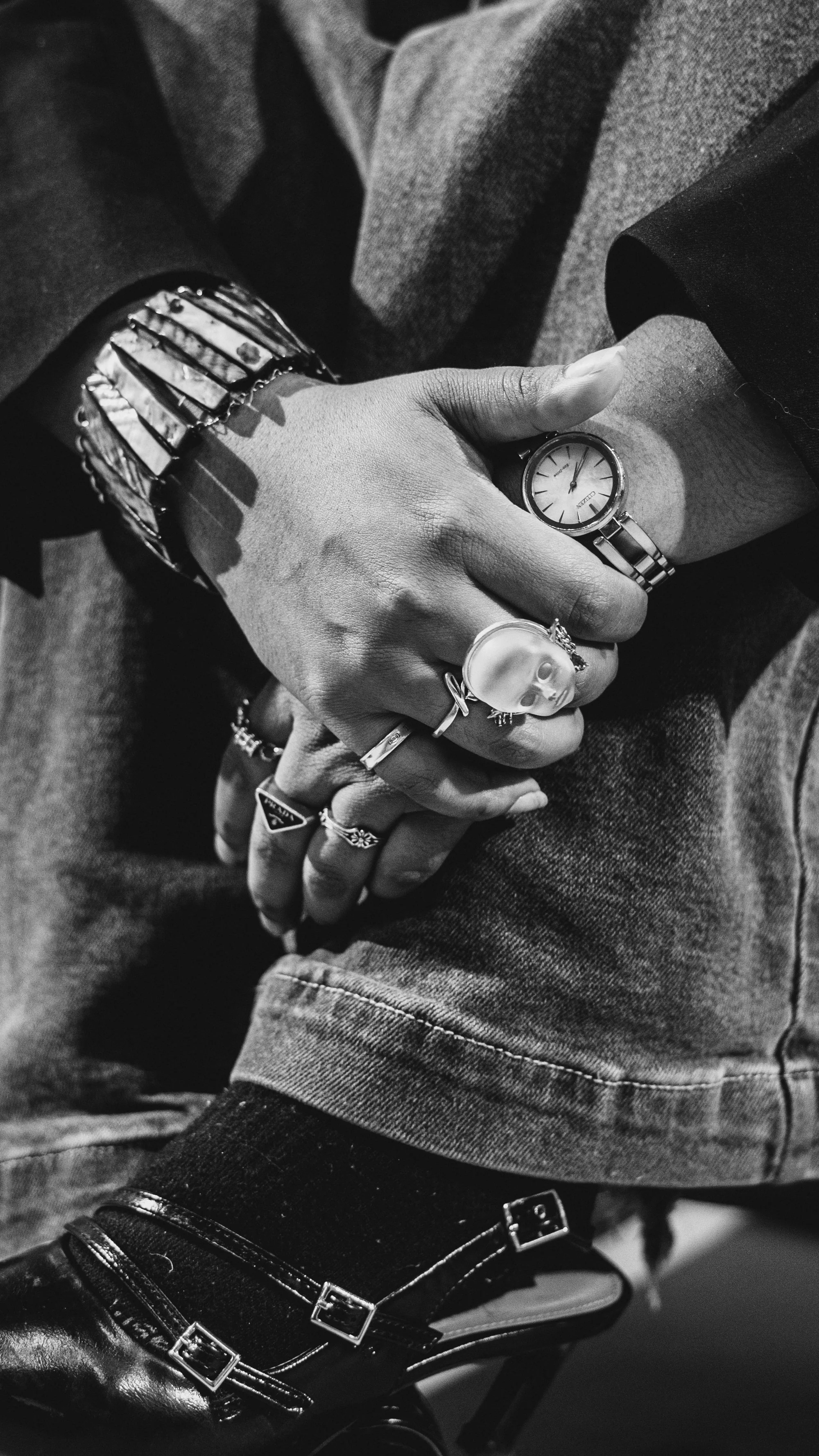 Close-up of a person's hand with multiple rings, wearing a wristwatch and bracelets, resting on their knee. The person is dressed in jeans and black shoes.