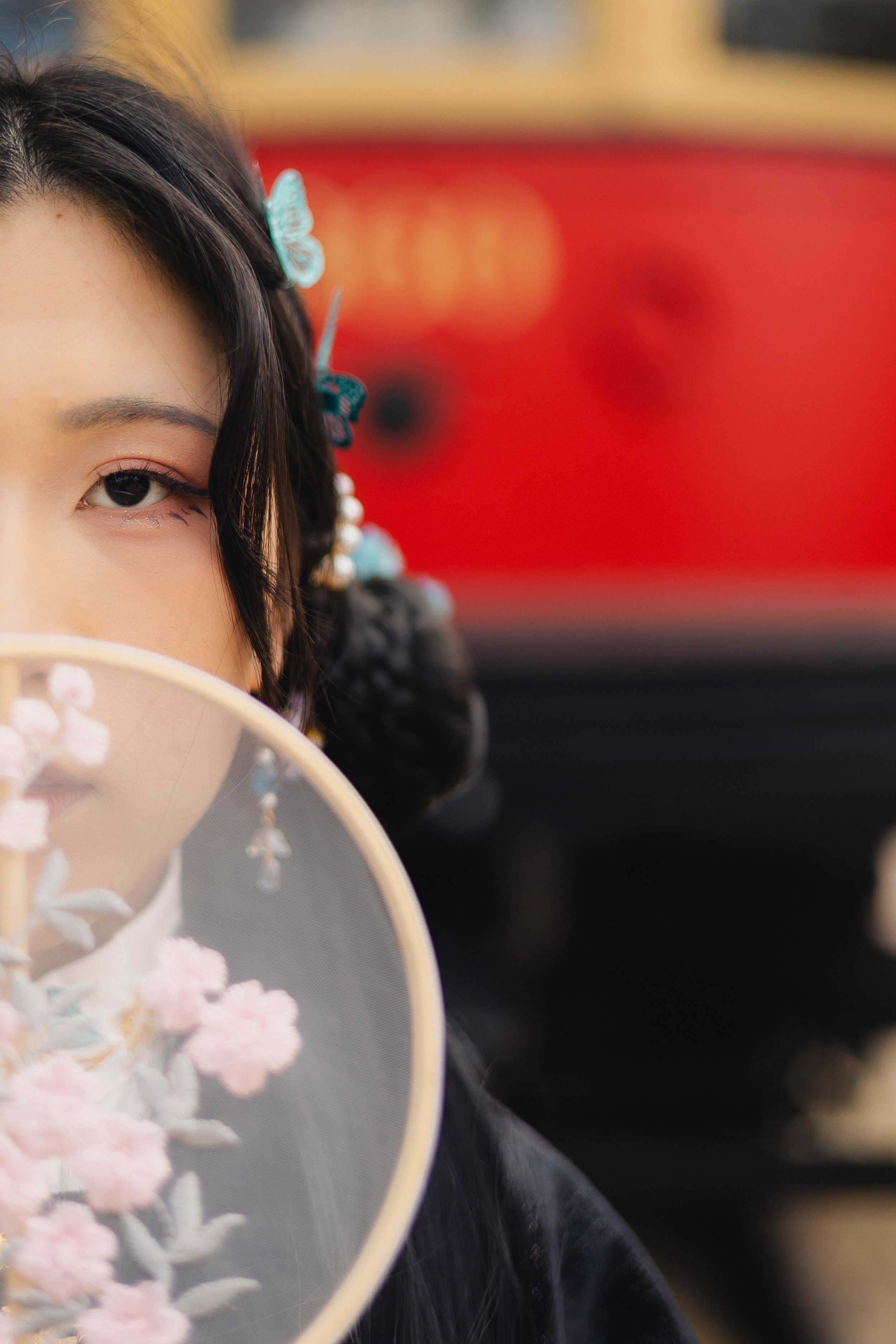 Close-up of a woman with dark hair decorated with blue butterfly hairpins, partially obscured by a hand fan with pink floral embroidery, standing outdoors with red and beige background.