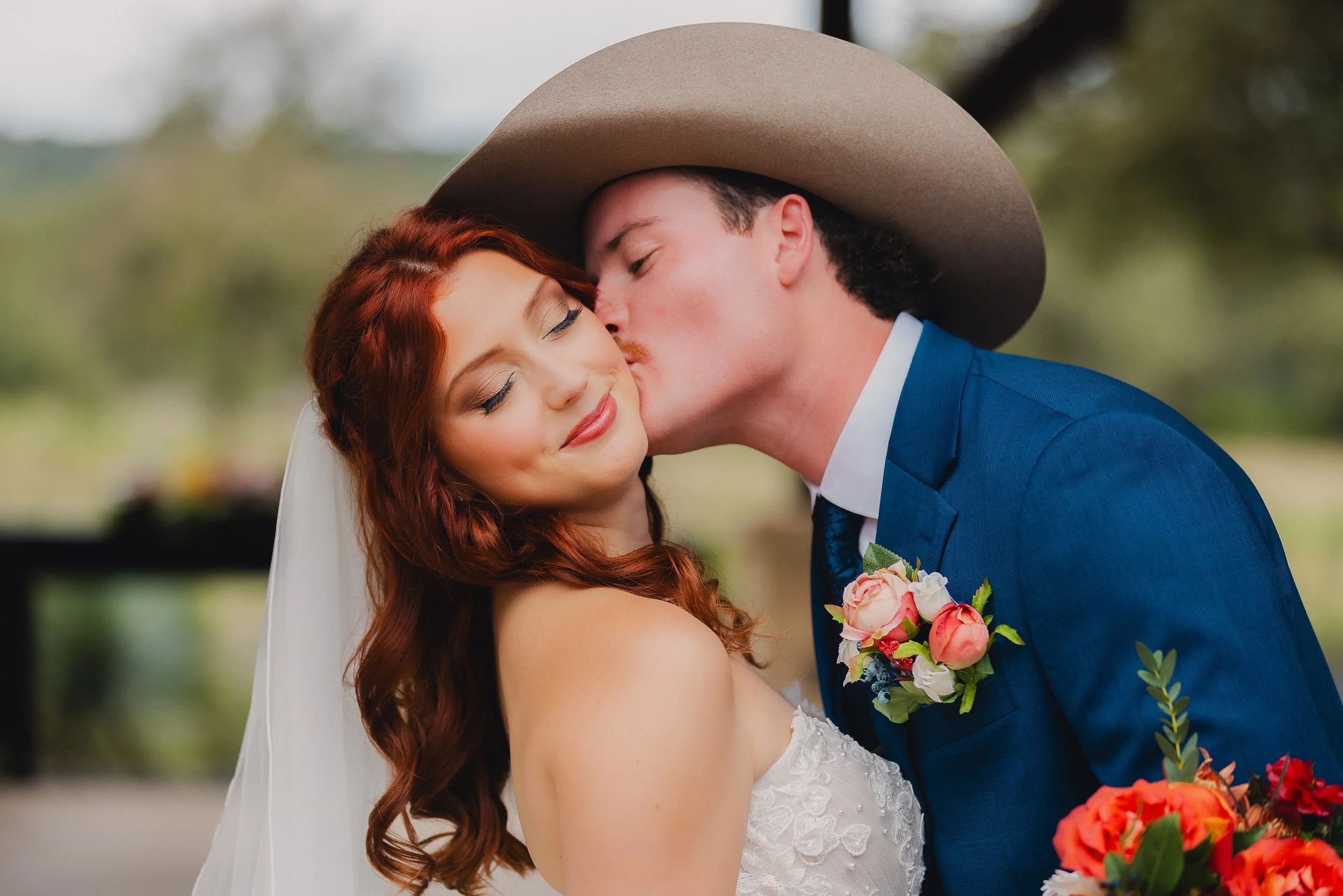 A bride with red hair and a groom in a blue suit sharing a romantic kiss outdoors; the groom is wearing a tan wide-brimmed hat, holding a bouquet of pink and white flowers.